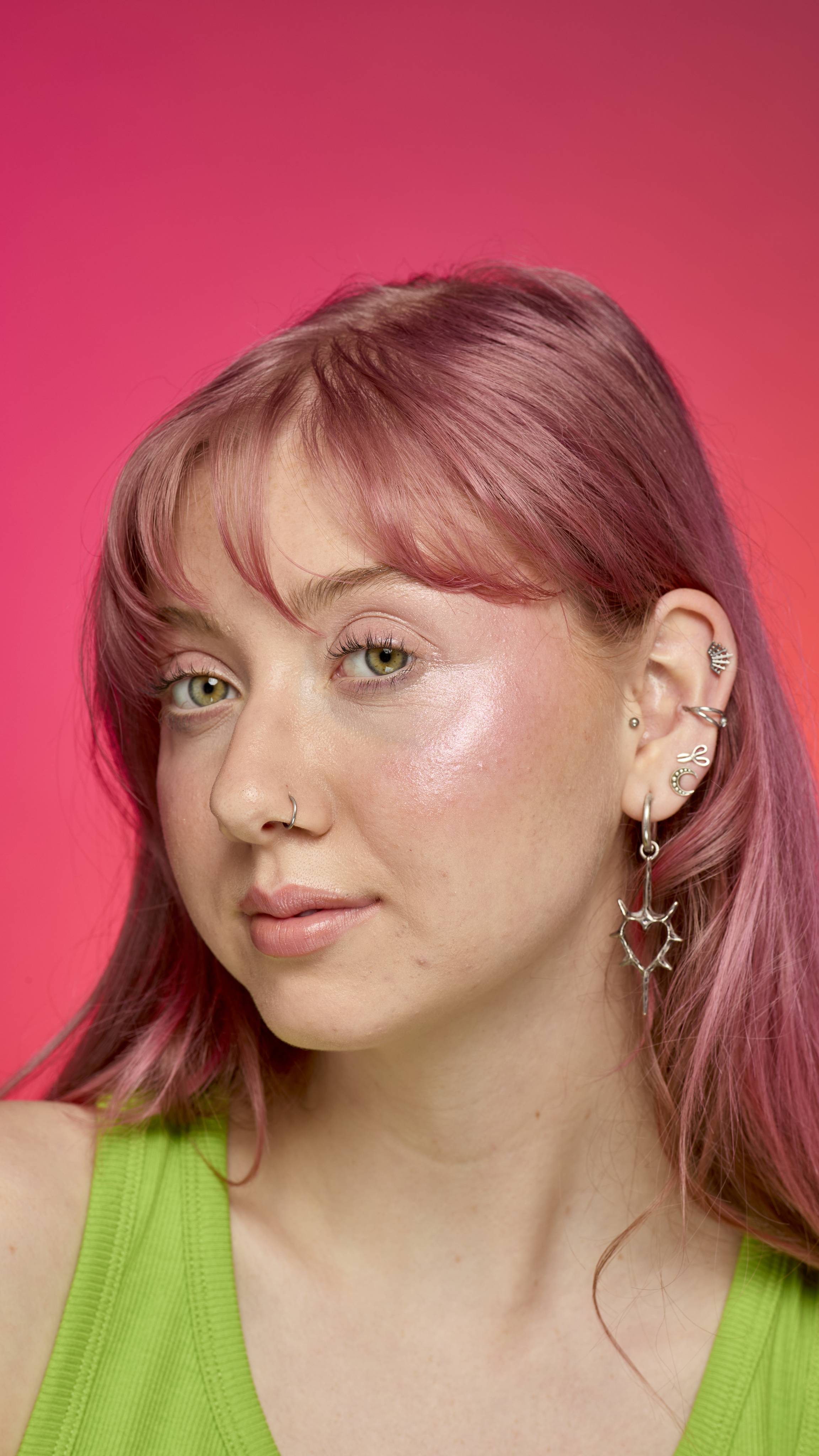 The image shows a model with pastel-pink hair on a hot pink background. They are wearing a green tank top as they look directly at the camera. Their cheeks have a shimmery, rosy glow from the Pink Glow Stick highlighter. 