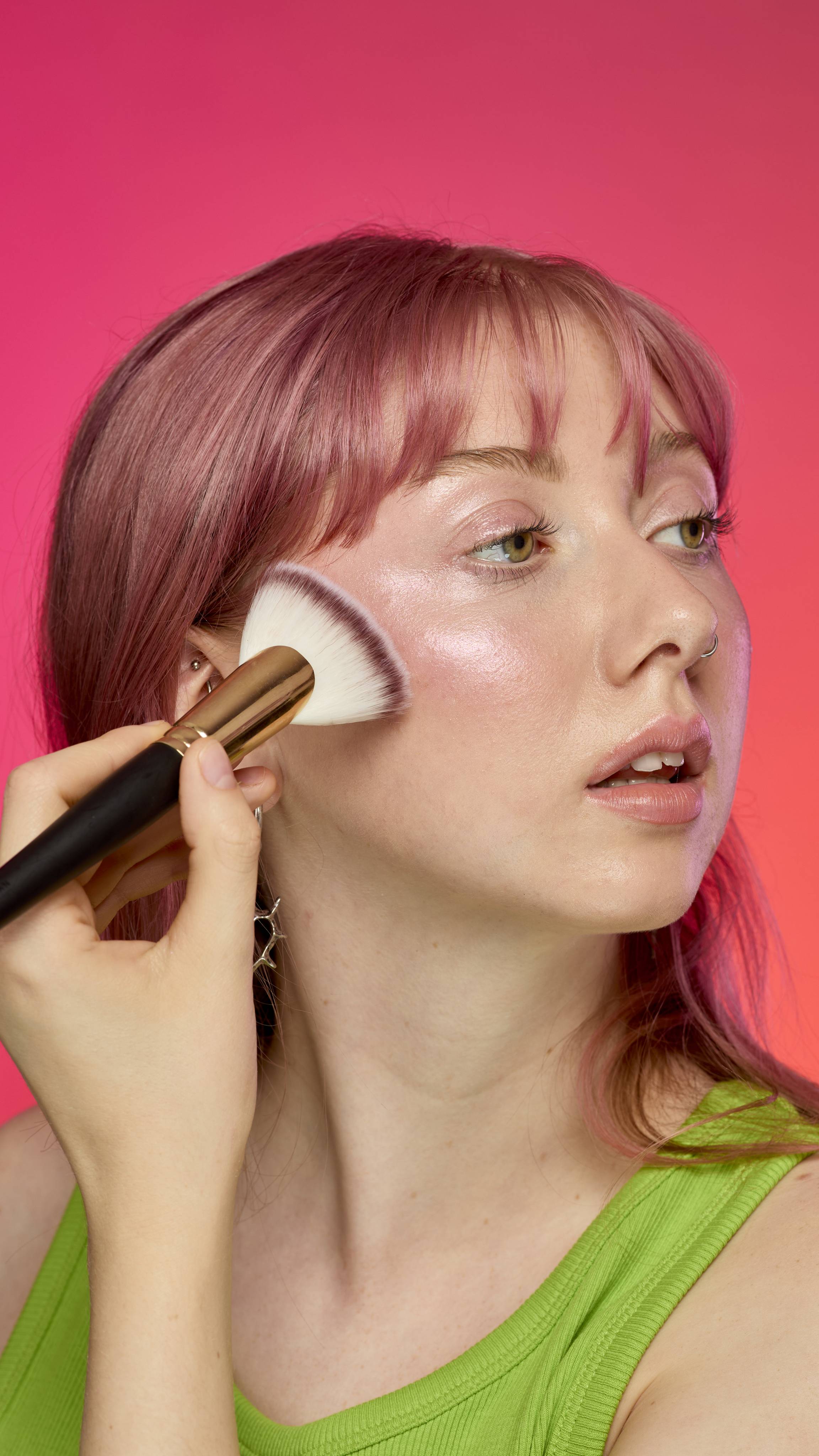 The image shows a model with pastel-pink hair on a hot pink background. They are wearing a green tank top as they apply the Pink Glow Stick highlighter to their cheekbones with a fanned brush. 