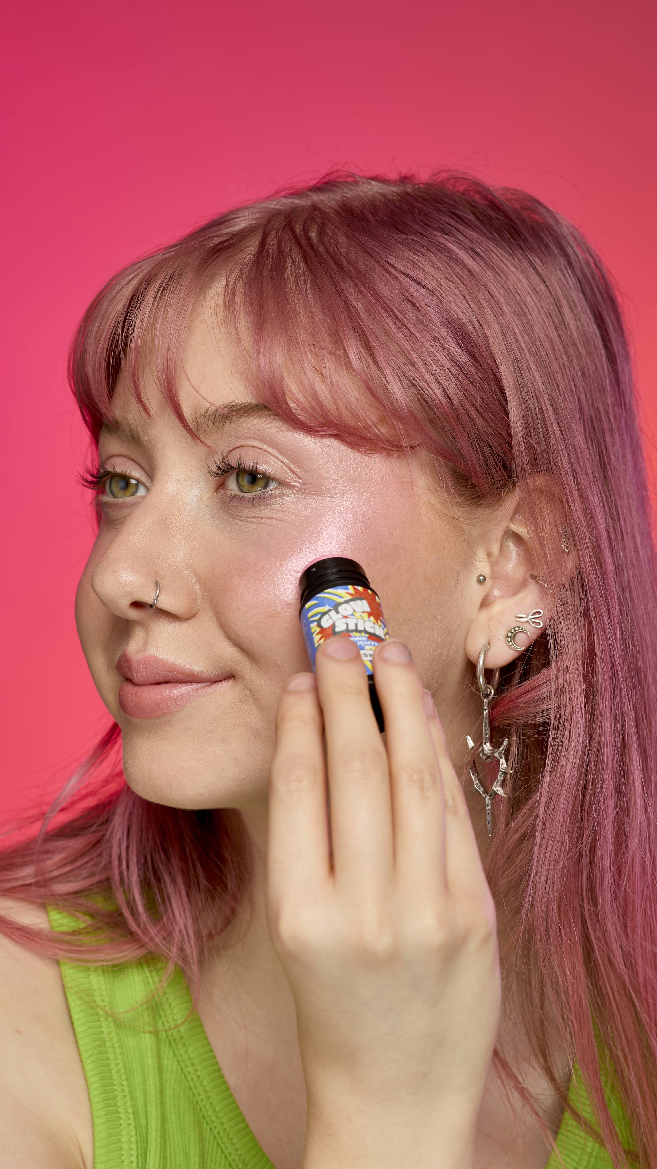 The image shows a model with pastel-pink hair on a hot pink background. They are wearing a green tank top as they apply the Pink Glow Stick highlighter to their cheekbones directly from the product applicator.