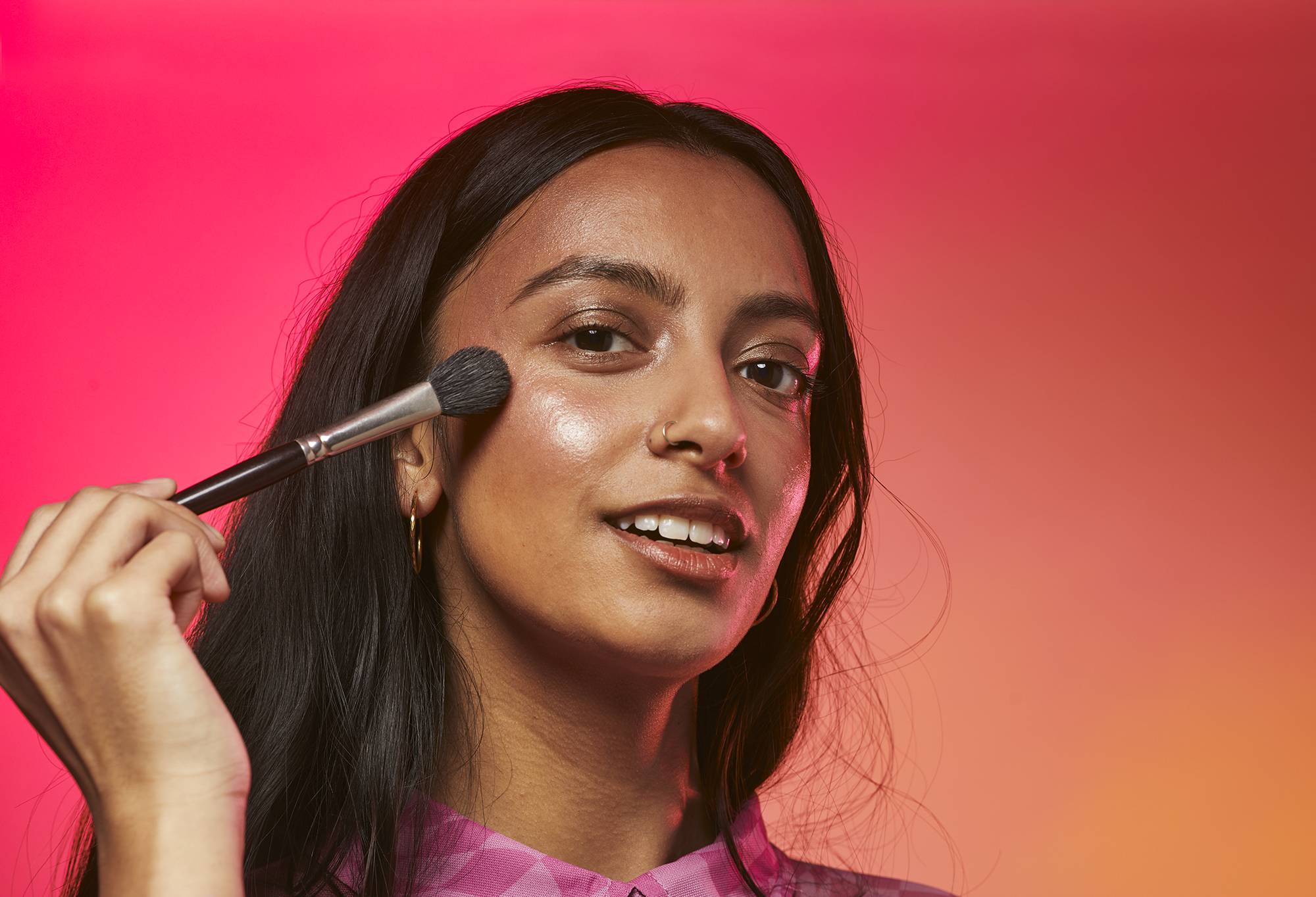A person smiles as they apply Pipit Glow Stick, a shimmery, rose gold highlighter, to their cheeks with a makeup brush.