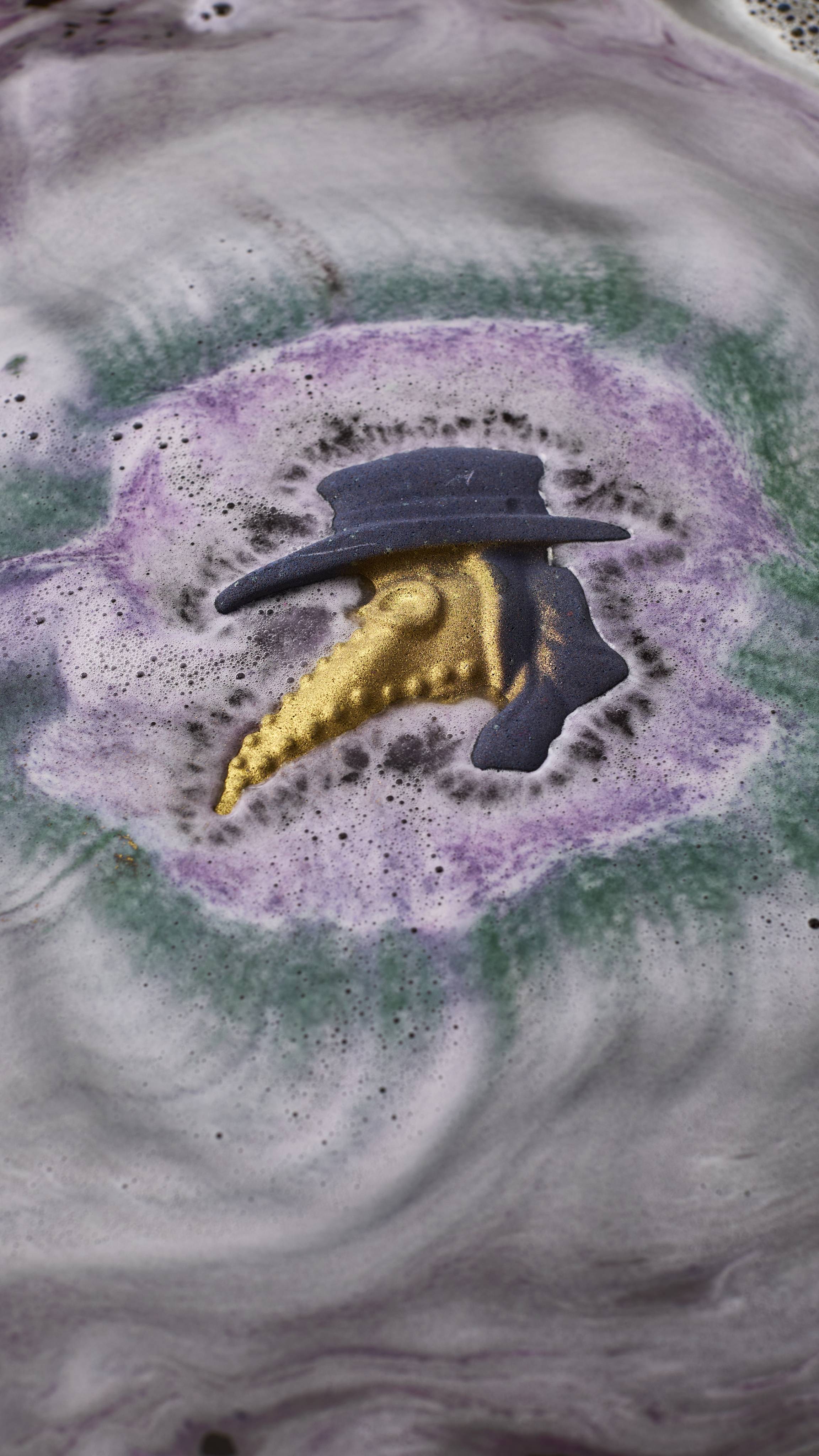 A plague doctor-shaped bath bomb dissolves in water, creating concentric ripples of purple, green, and dark grey froth around its purple hat and shimmering gold-beaked mask. Swirling patterns of light grey foam cover the rest of the bath.