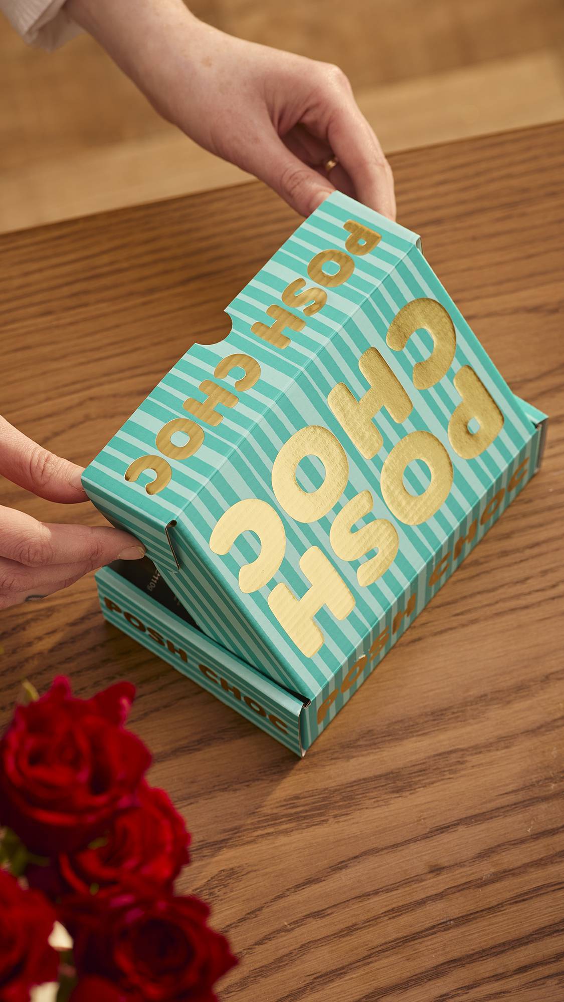 The image shows the Posh Choc, teal-striped gift box on a wooden counter with red roses just visible at the bottom. The model's hand is mid-way through opening the gift box. 