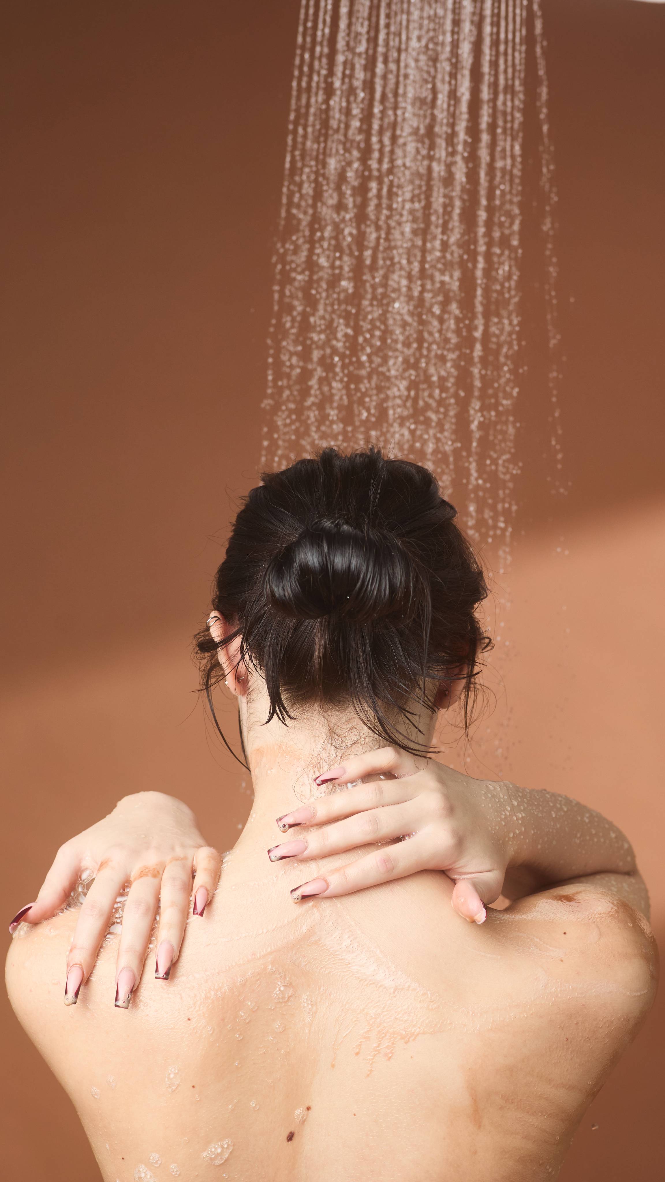 The model is standing under running shower water on a warm, earth-toned background as they lather their neck and shoulder with the Posh Chocolate shower gel.