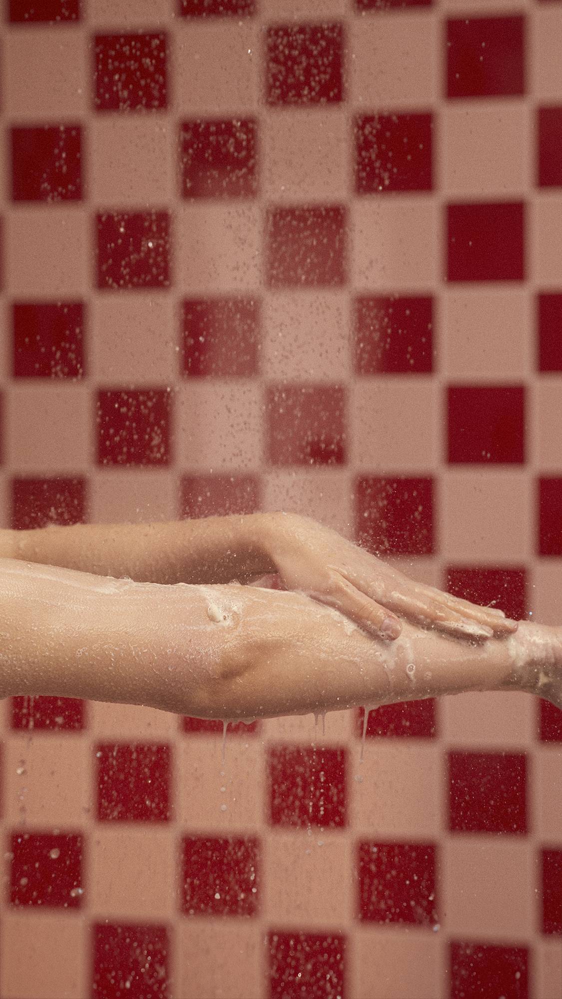 A close-up image of the model's arm under shower water as they soothe the body wash over their forearm. 