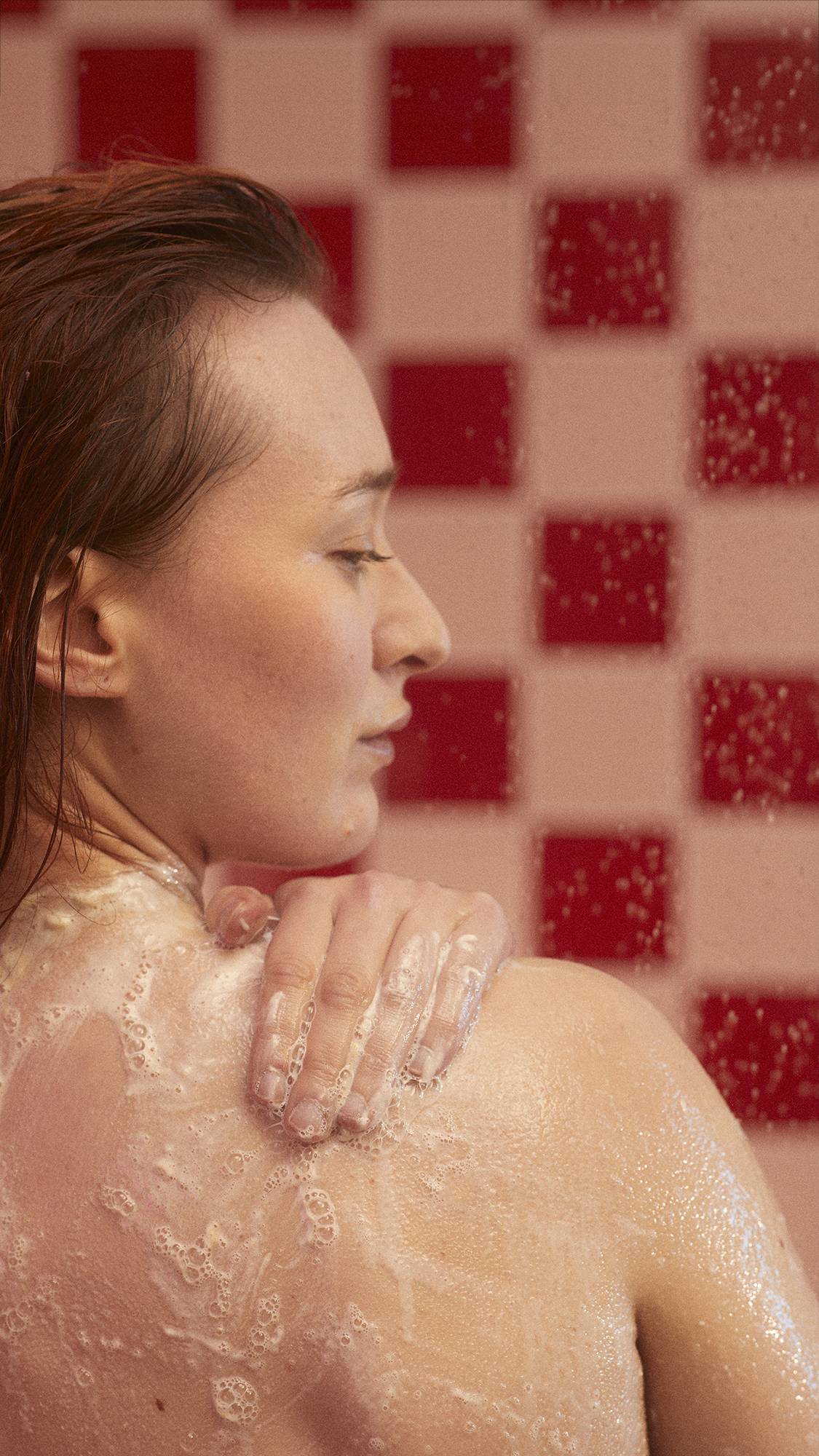 The model is in a tiled backroom under shower water as they lather their shoulder with Posh White Chocolate and Rose body wash. 