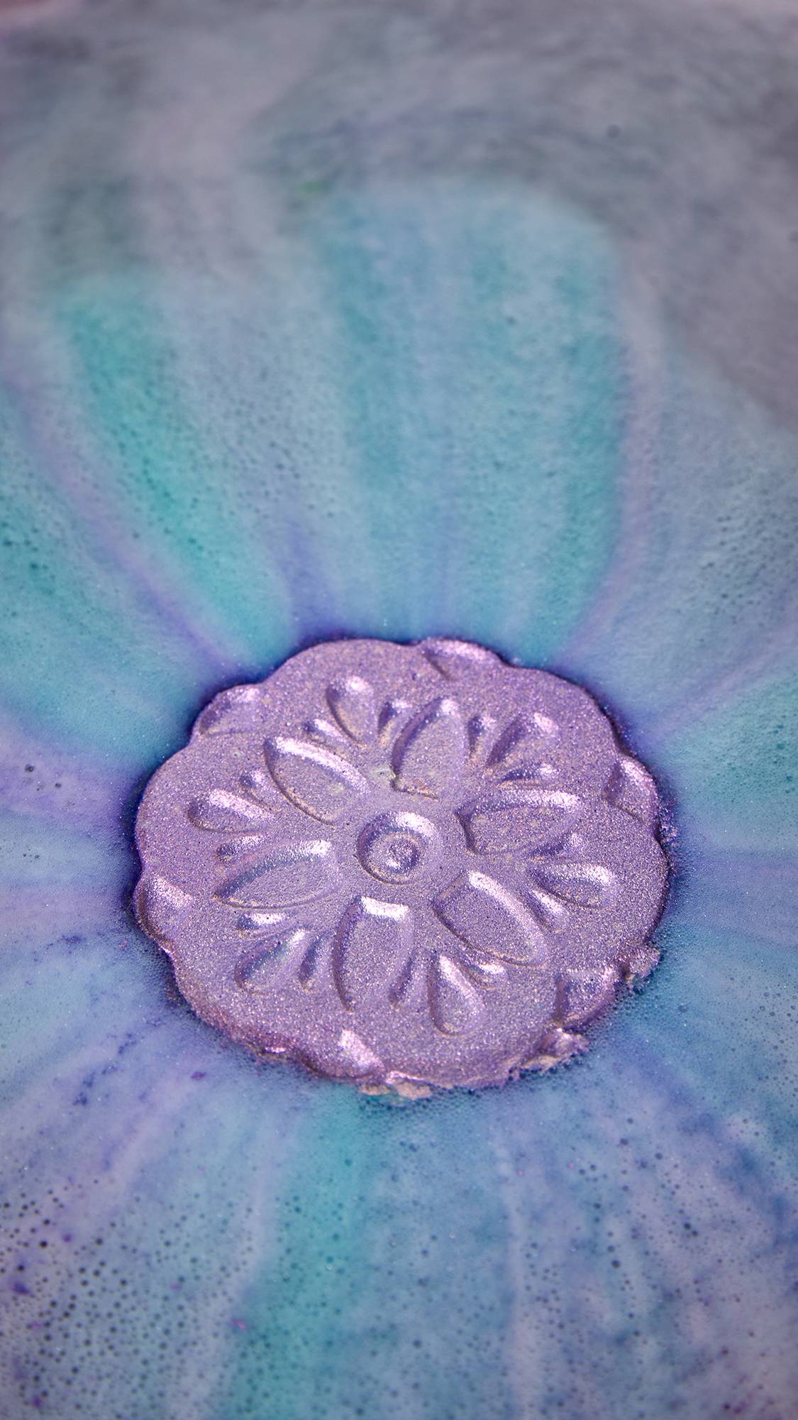 A shimmery purple, flower-shaped bath bomb floats in a bath of swirling blue and purple water. The water is covered in a delicate layer of bubbles, and the bomb glistens with plastic-free glitter.