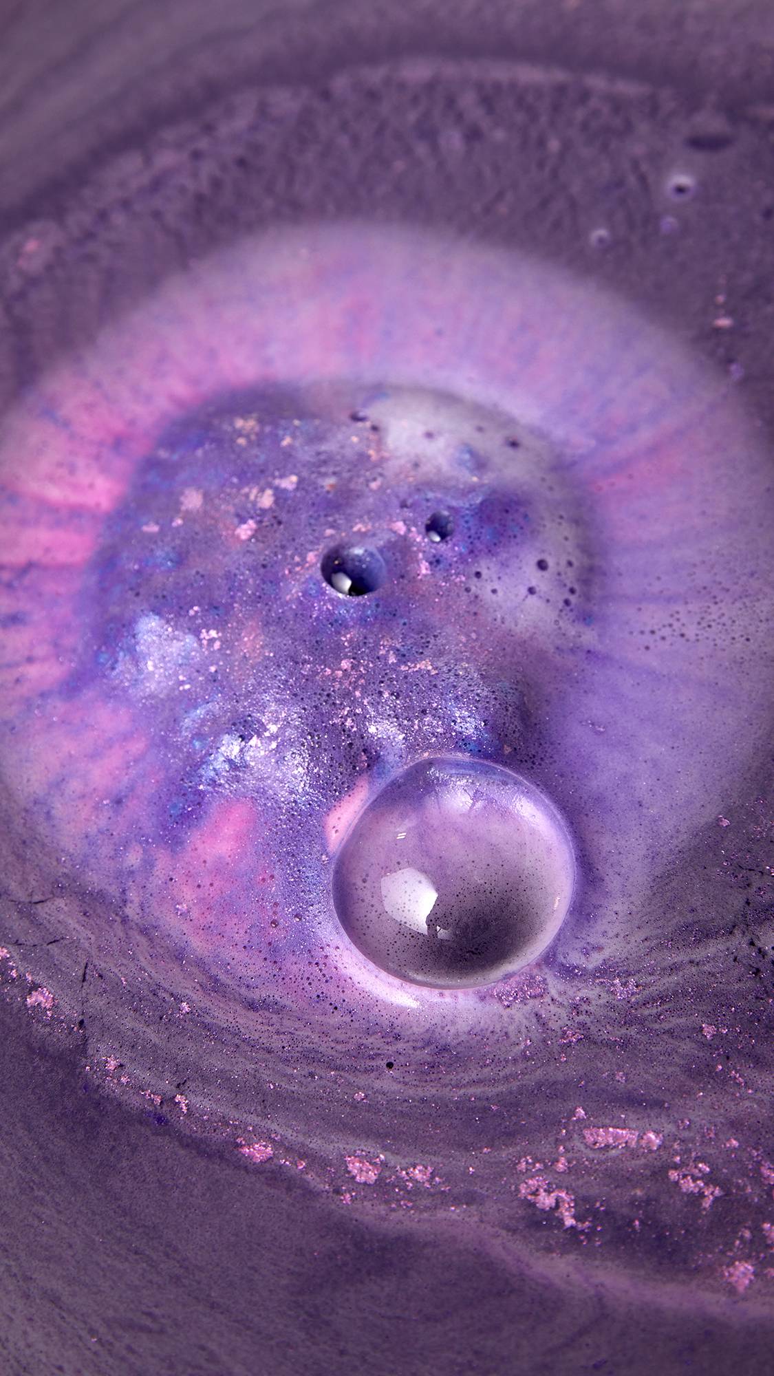 A bath bomb dissolves in purple water, releasing a shimmering pink and purple fizz. A large, clear bubble floats on the surface of the foamy water, which sparkles with plastic-free glitter.