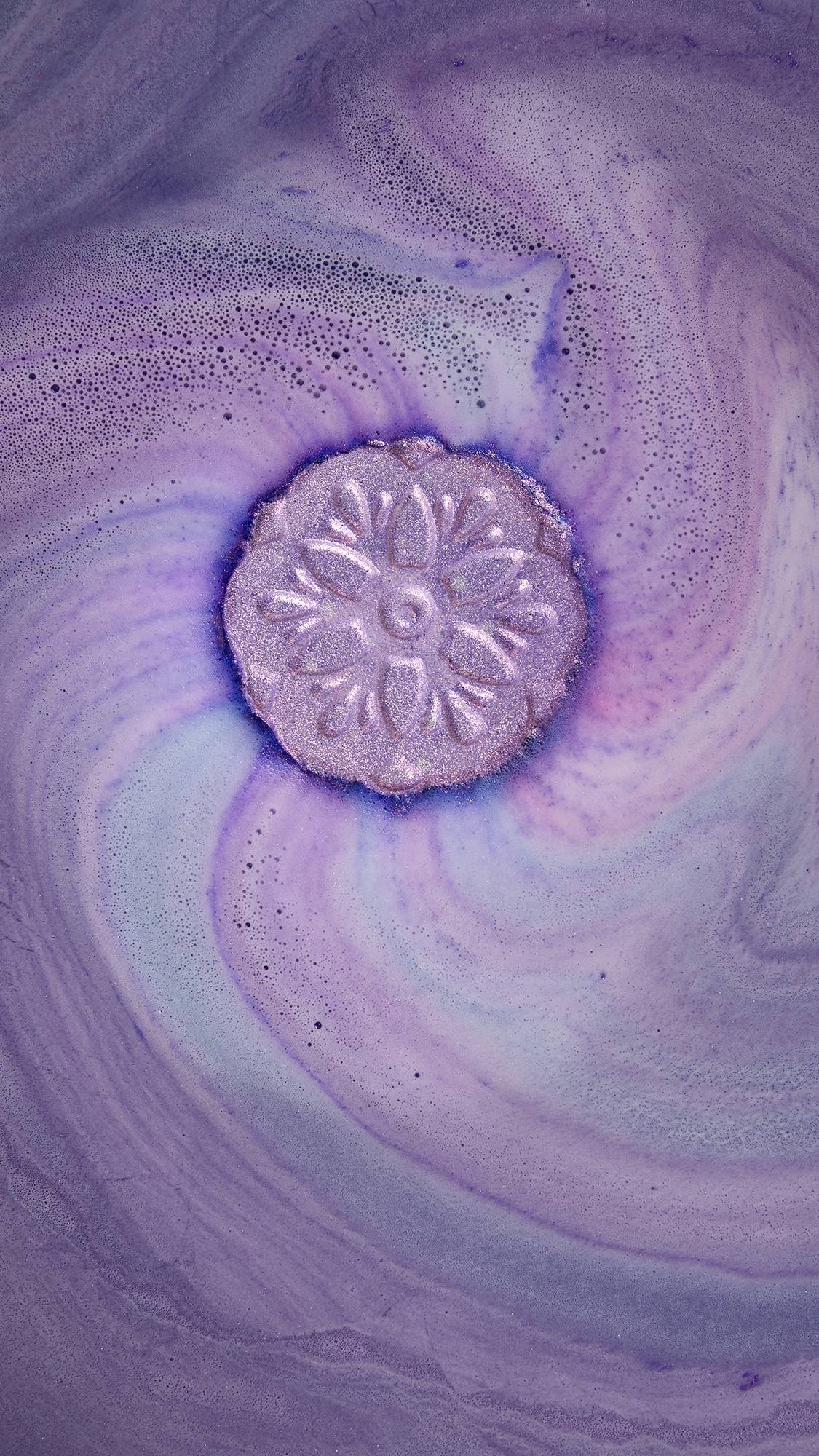 A close-up of a shimmering purple bath bomb with a flower design, fizzing and creating a psychedelic spiral of purple and white foam. The mesmerizing, vibrant shimmer is from plastic-free glitter.