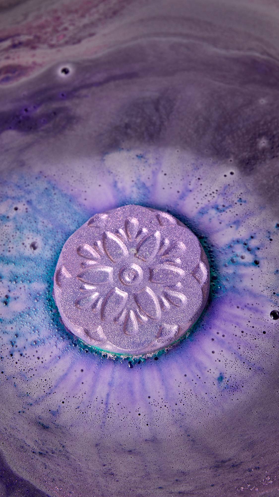 A sparkling purple bath bomb with a flower design fizzes in a pool of swirling purple and blue water, creating foam. The bath bomb shimmers with plastic-free glitter.