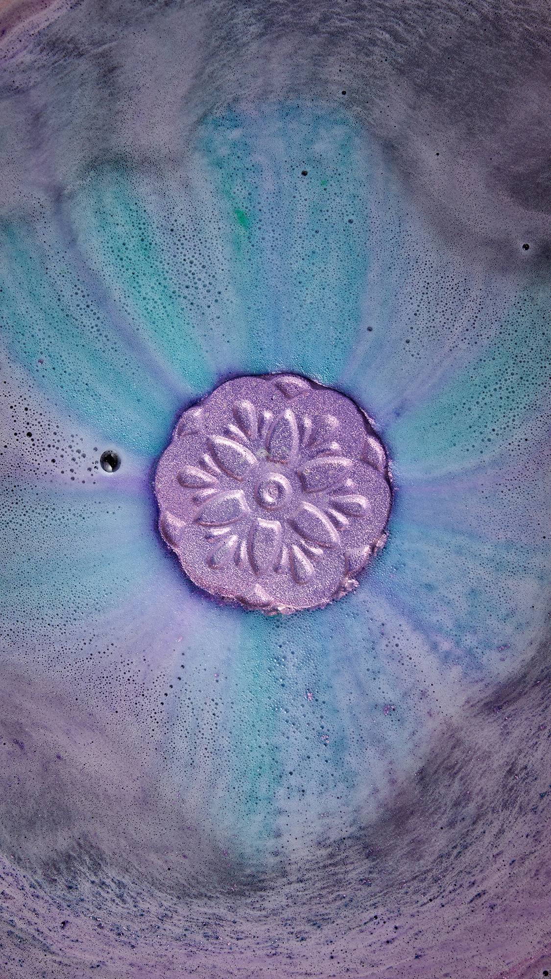 A mesmerizing purple bath bomb with a flower design fizzes, releasing a swirl of turquoise and violet hues into the water. The shimmering effect comes from our plastic-free glitter, offering a guilt-free, colorful soak.