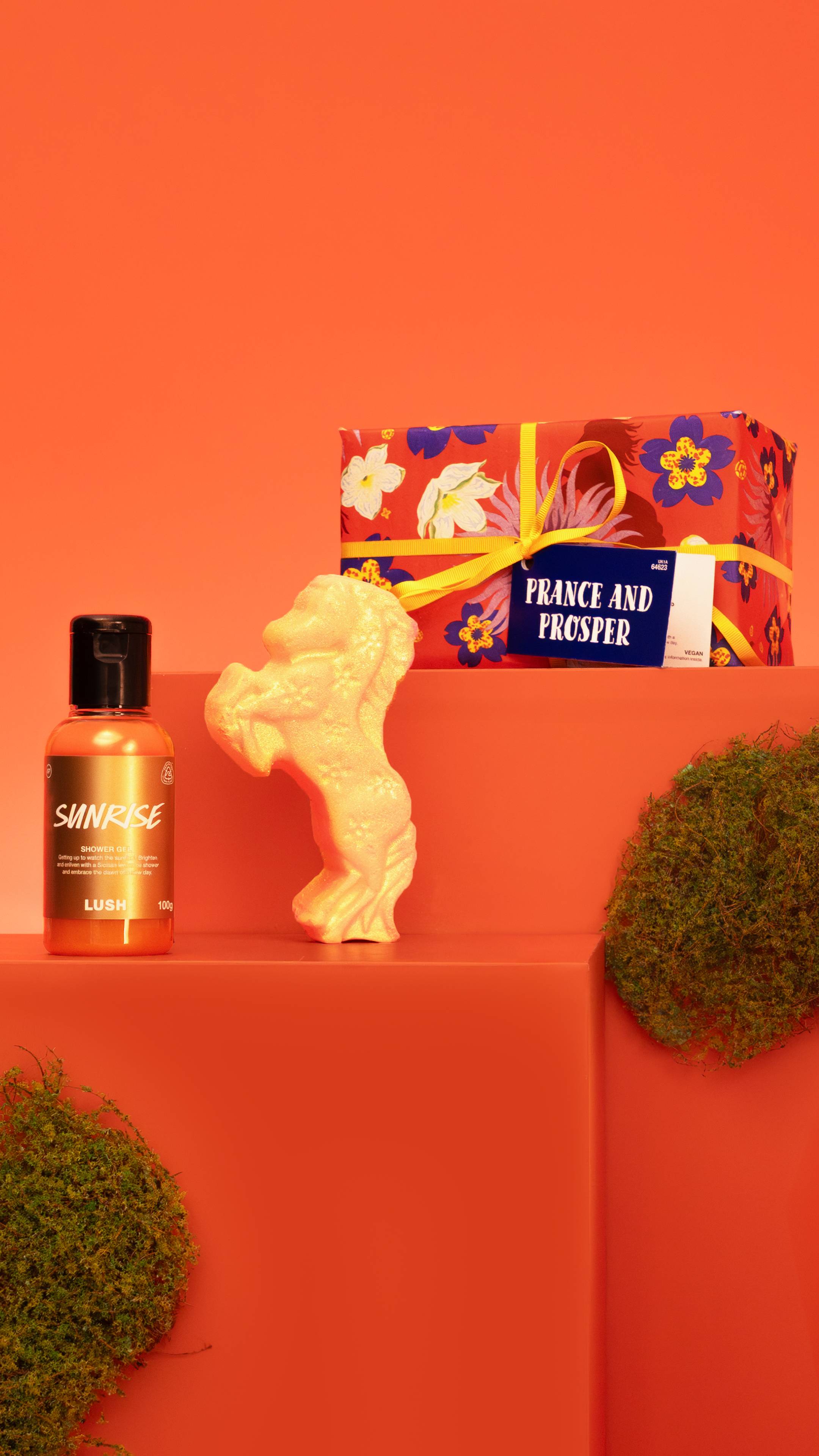 The Prance and Prosper gift set, wrapped in red floral paper, stands next to a bottle of Sunrise shower gel and a gold horse bath bomb shimmering with Plastic Free Glitter on an orange surface with moss.