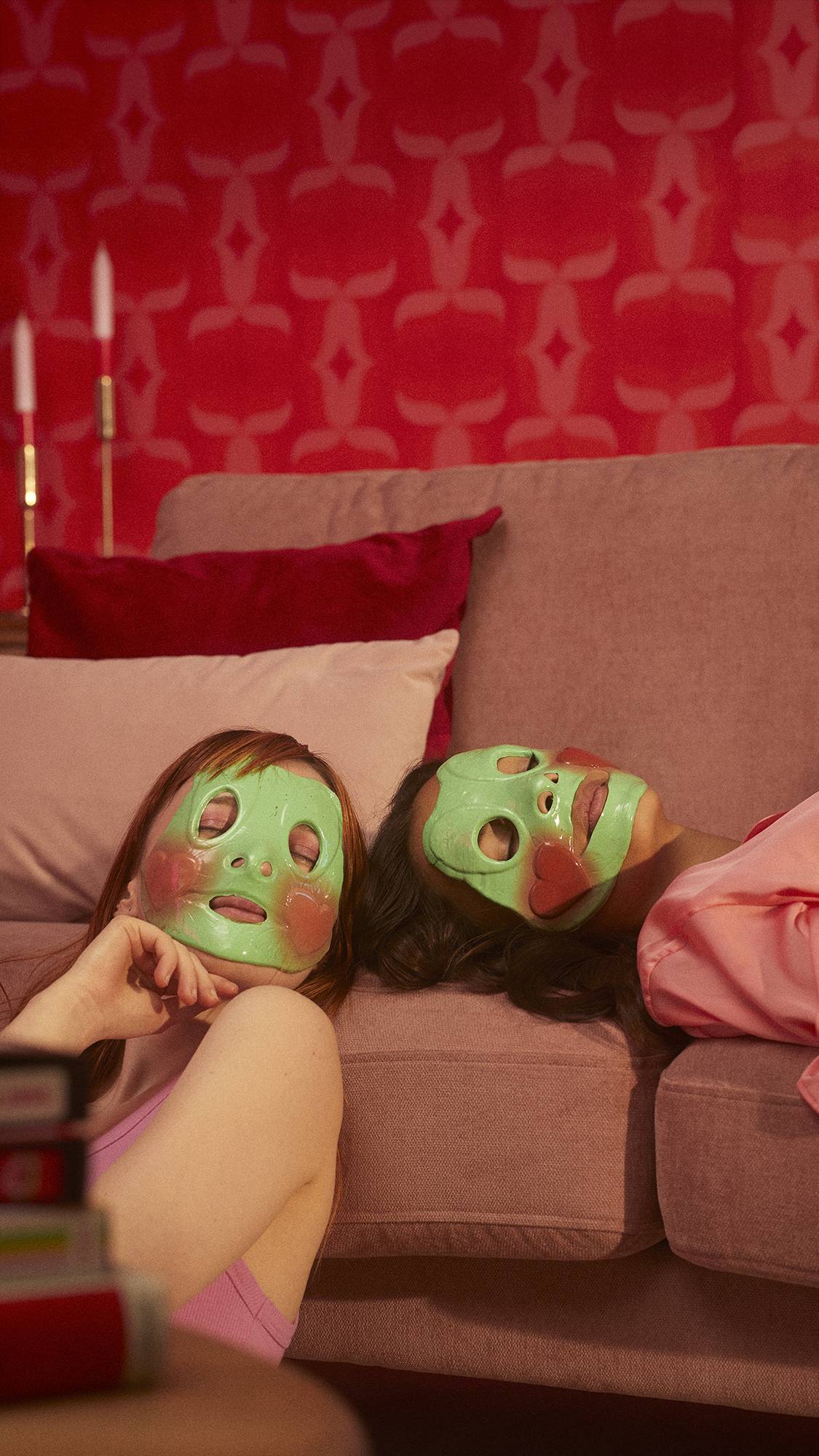 Two models are in a red room lying against a comfy sofa. They are both wearing the Prince Charming face mask. 