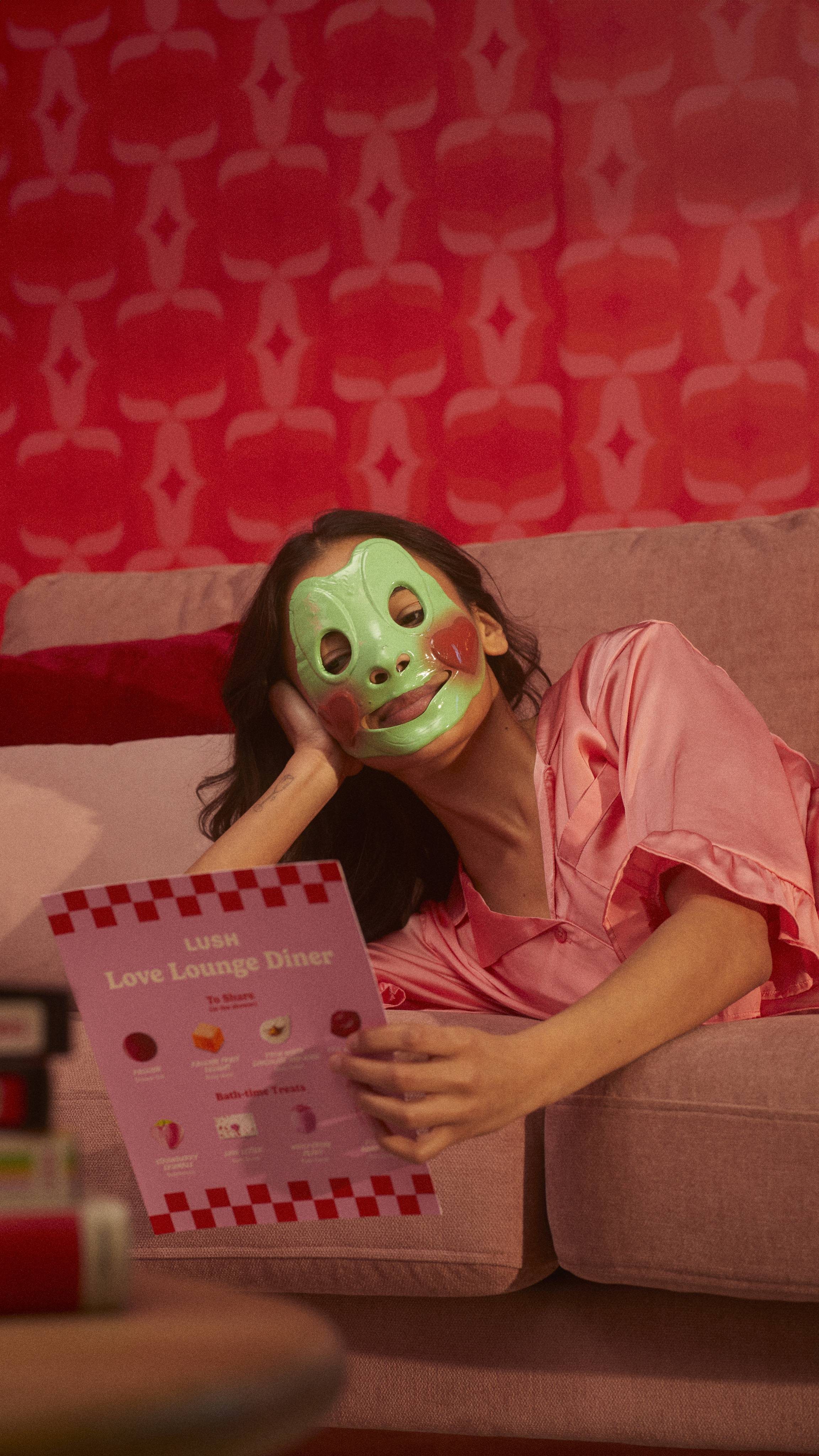 The model is laid down on a sofa in pink satin pyjamas reading a leaflet with the Prince Charming face mask on.