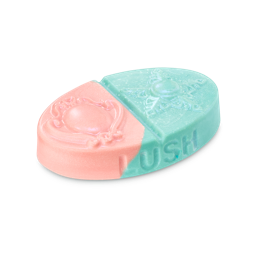 Oval body balm split diagonally into pink and teal sections with embossed brooch and star designs. Features the 'LUSH' logo on the side  with a fine shimmer.