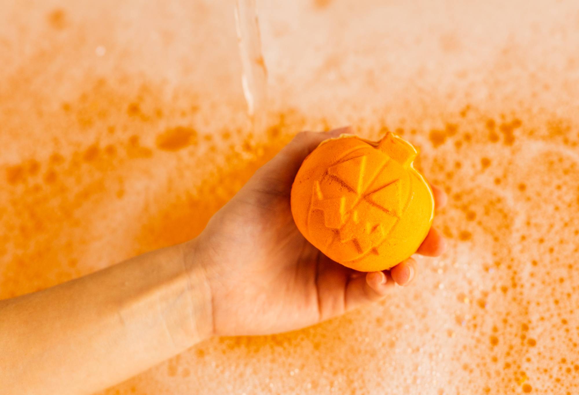 Image shows model holding the bubble bar over a thick blanket of foamy bubbles and bright orange bath water.