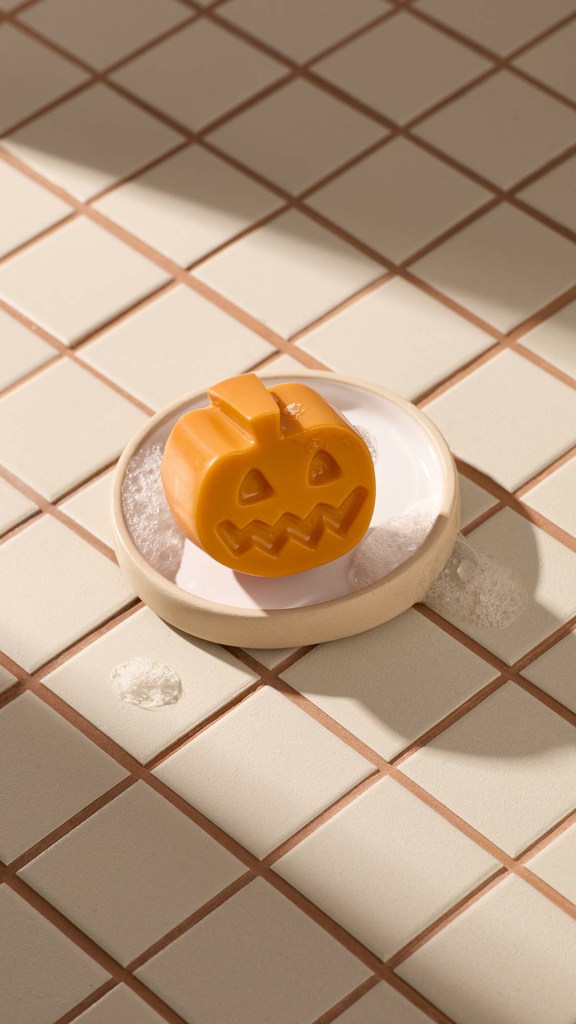 An orange Lush pumpkin facial gummy sits in a round beige soap dish, surrounded by white soapy foam. The dish rests on light beige square tiles with brown grouting. Diagonal sunny shadows cross the scene.