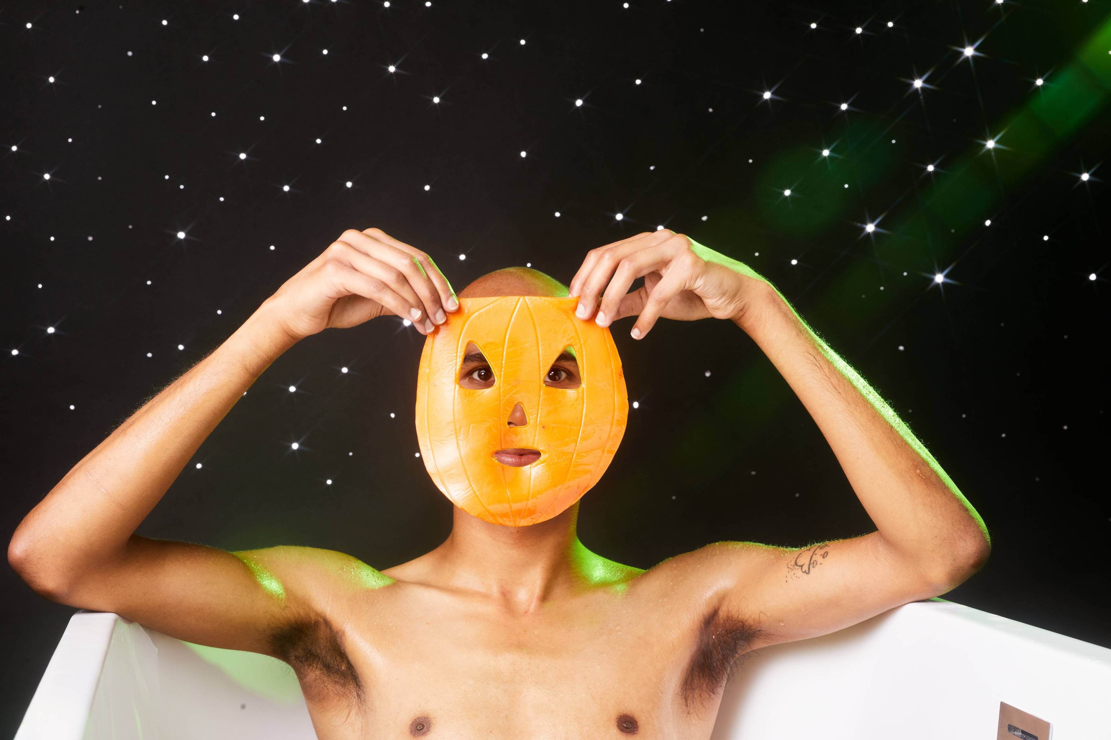 Image shows the model sitting in a bath on a twinkling background. They are holding the orange sheet mask up to their face.
