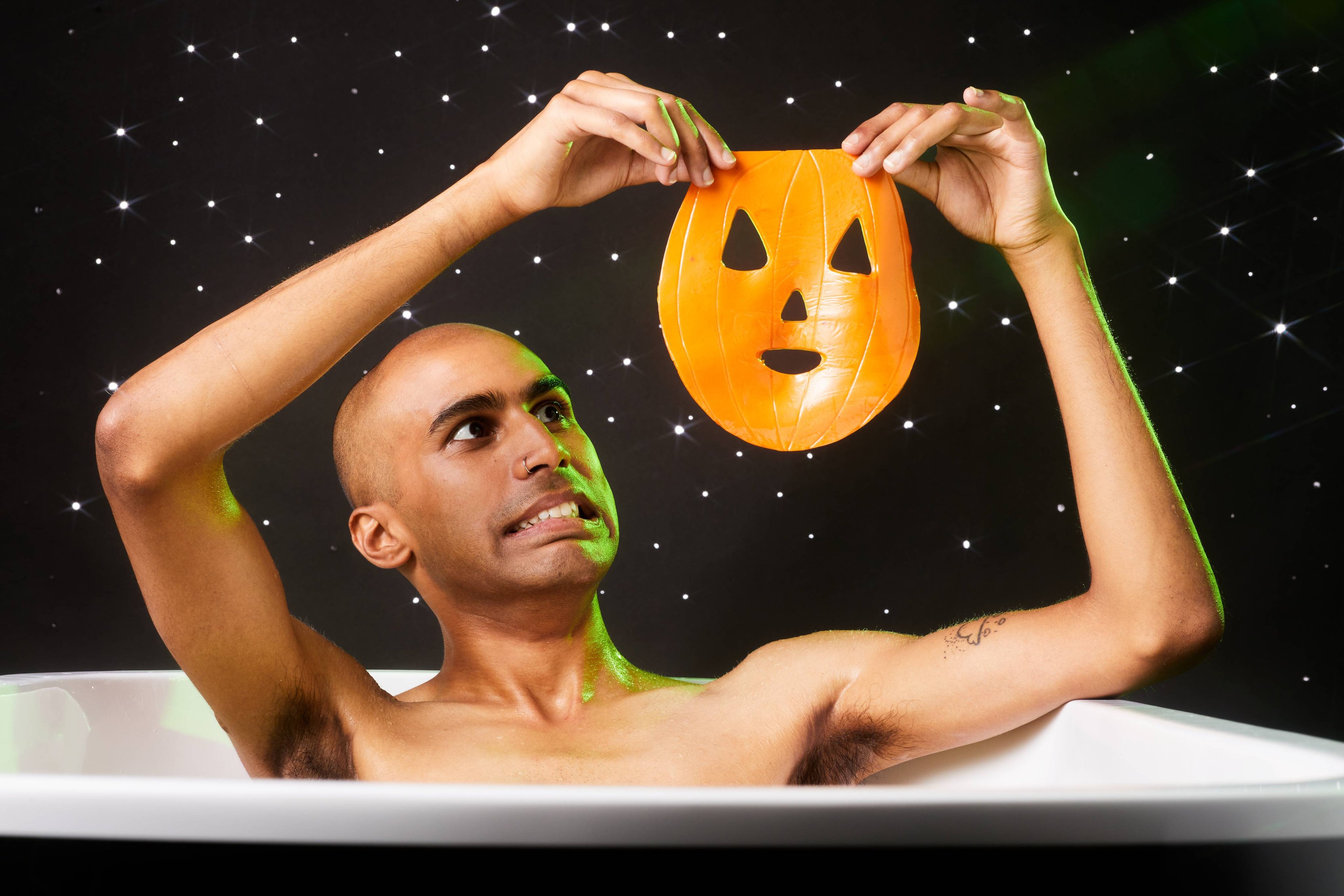 Image shows the model sitting in a bath on a twinkling background. They are holding the orange sheet mask up in the air.