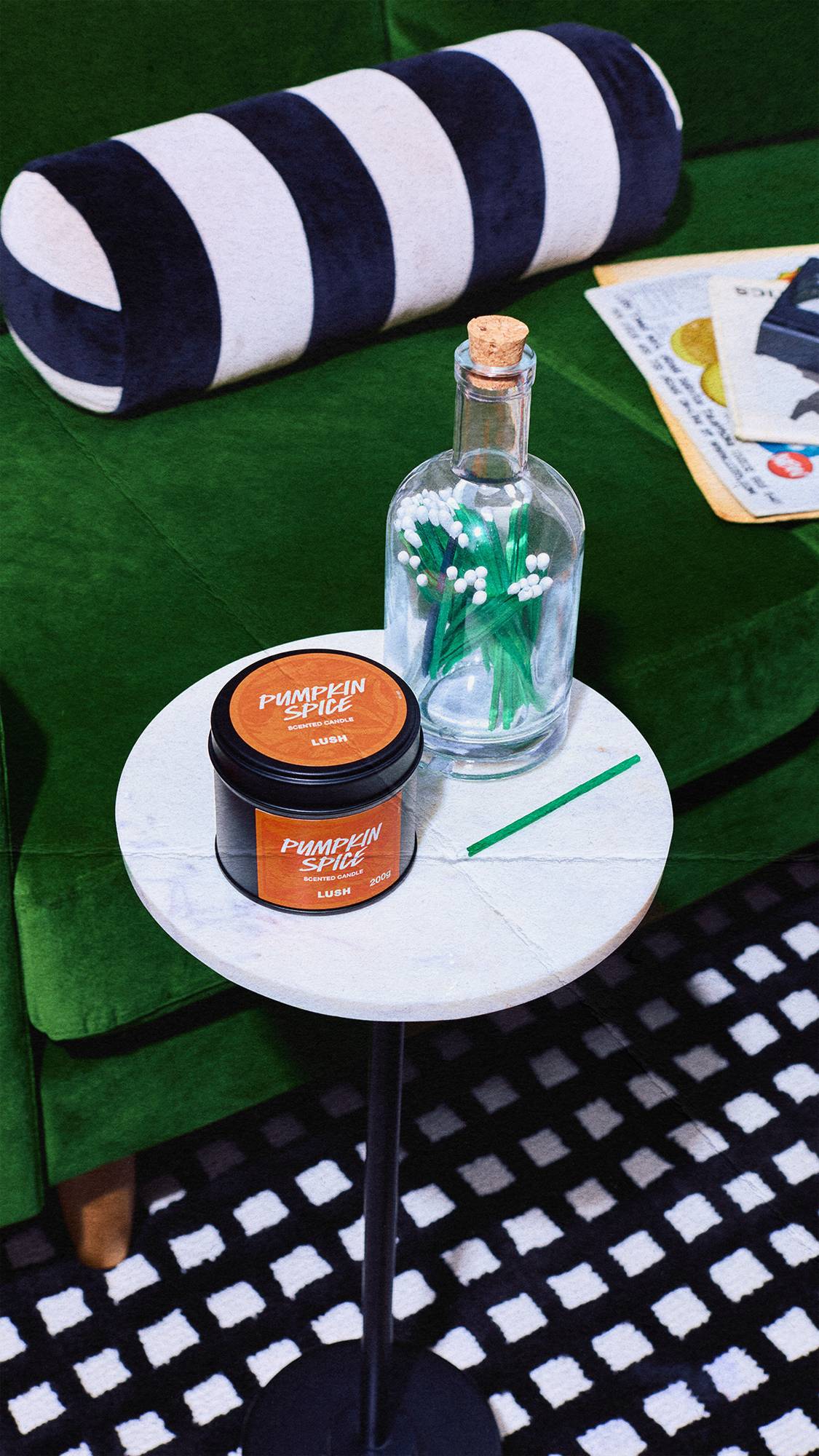 A Lush Pumpkin Spice scented candle and a bottle of green matches rest on a white marble side table, set against a dark green velvet sofa with a striped cushion.