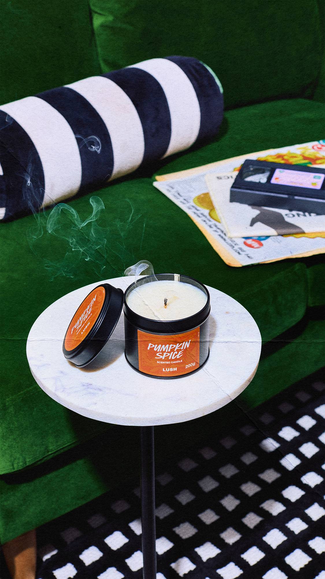 Wisps of smoke rise from a newly extinguished Lush Pumpkin Spice candle resting on a white marble table, set against a dark green velvet sofa with a striped cushion.