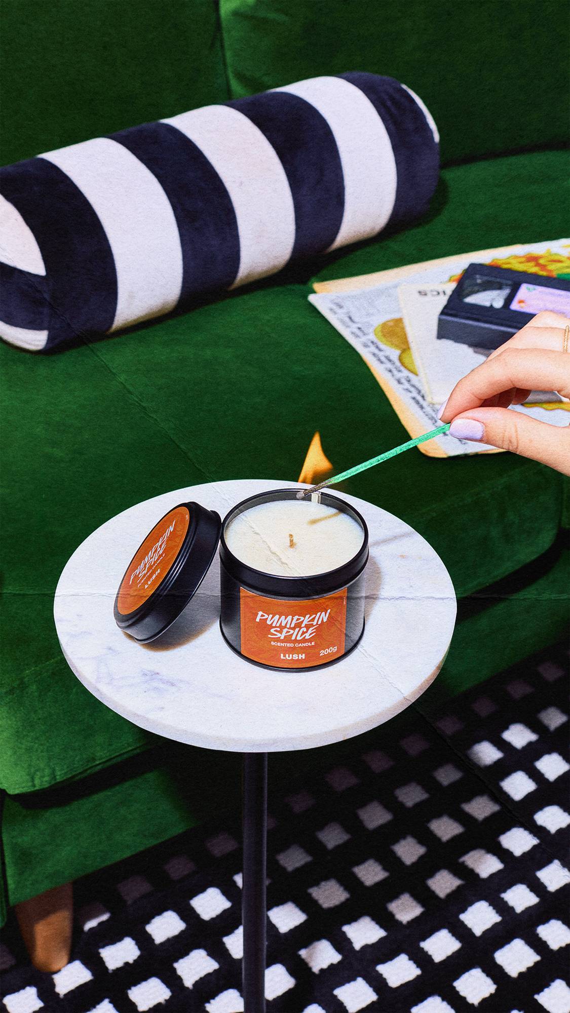 A hand holding a lit green match lights a open Lush Pumpkin Spice scented candle on a white marble table, set in a cosy room with a dark green velvet sofa.