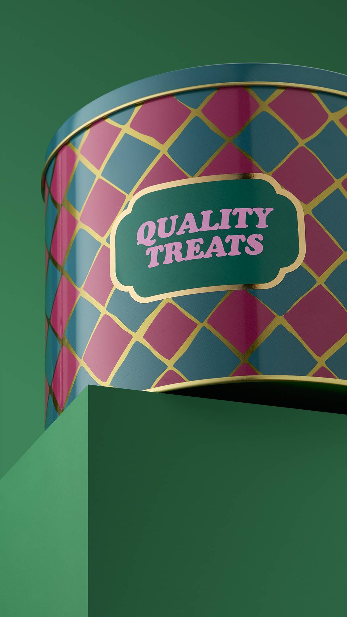 The image shows the colourful, cylindrical "Quality Treats" metal gifting tin, with a pink and teal checkered pattern, sitting on a green platform against a matching green background. 