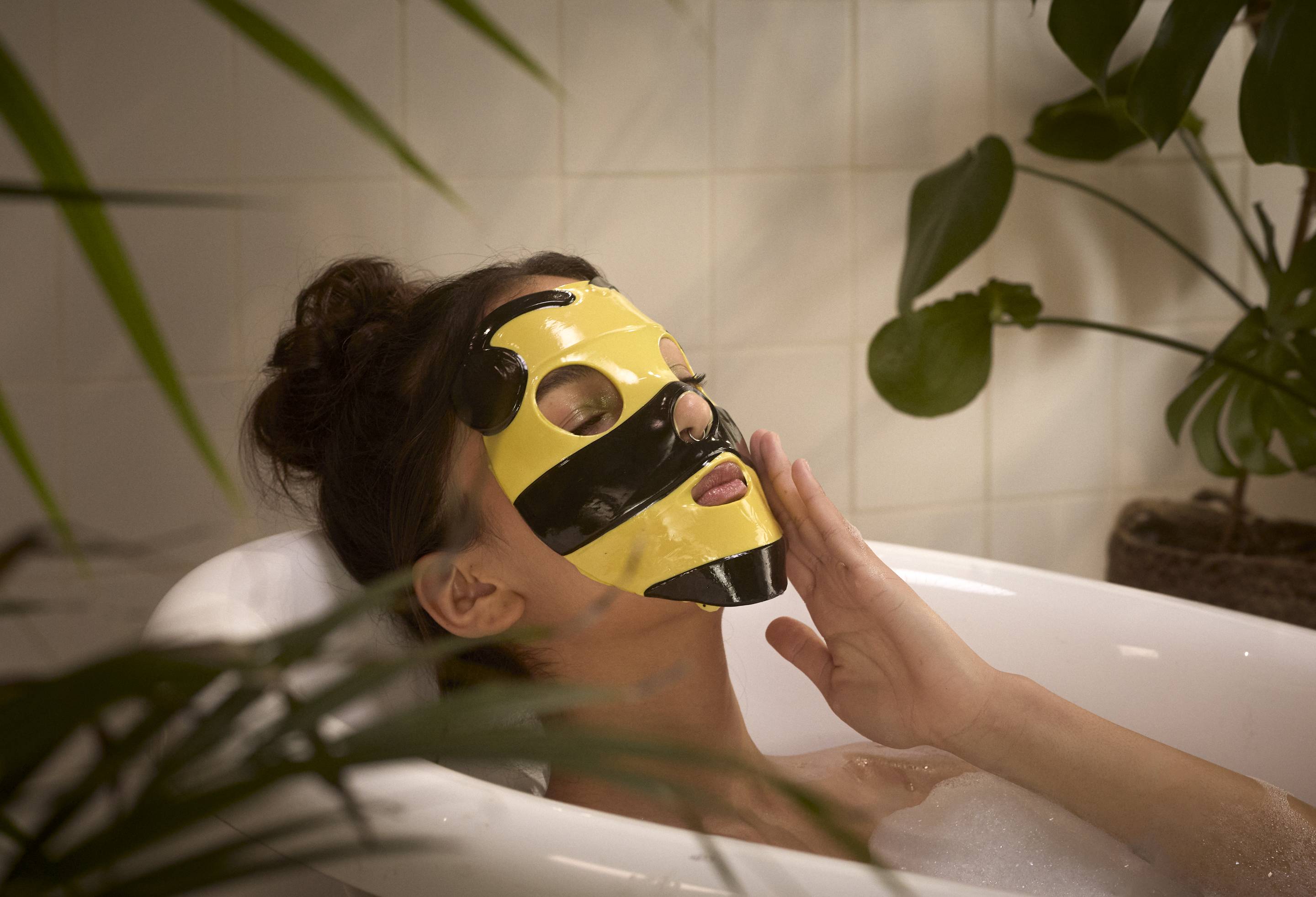 The image shows the model laid back in the bathtub with the Queen Bee sheet mask laid over their face.
