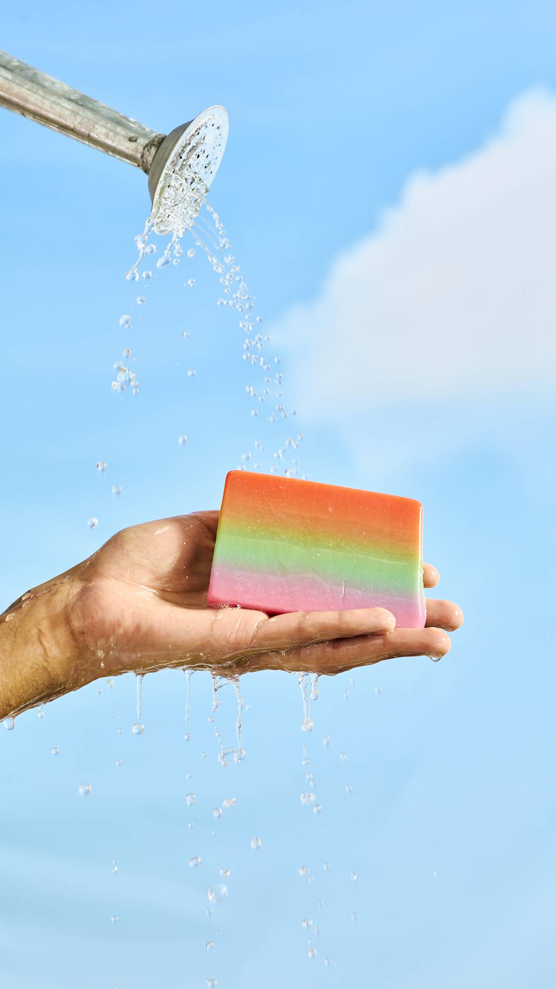A close-up of the model's flat hand holding the Rainbows and Waterfalls soap in their palm. There are blue skies and a watering can head is pouring water over the hand. 