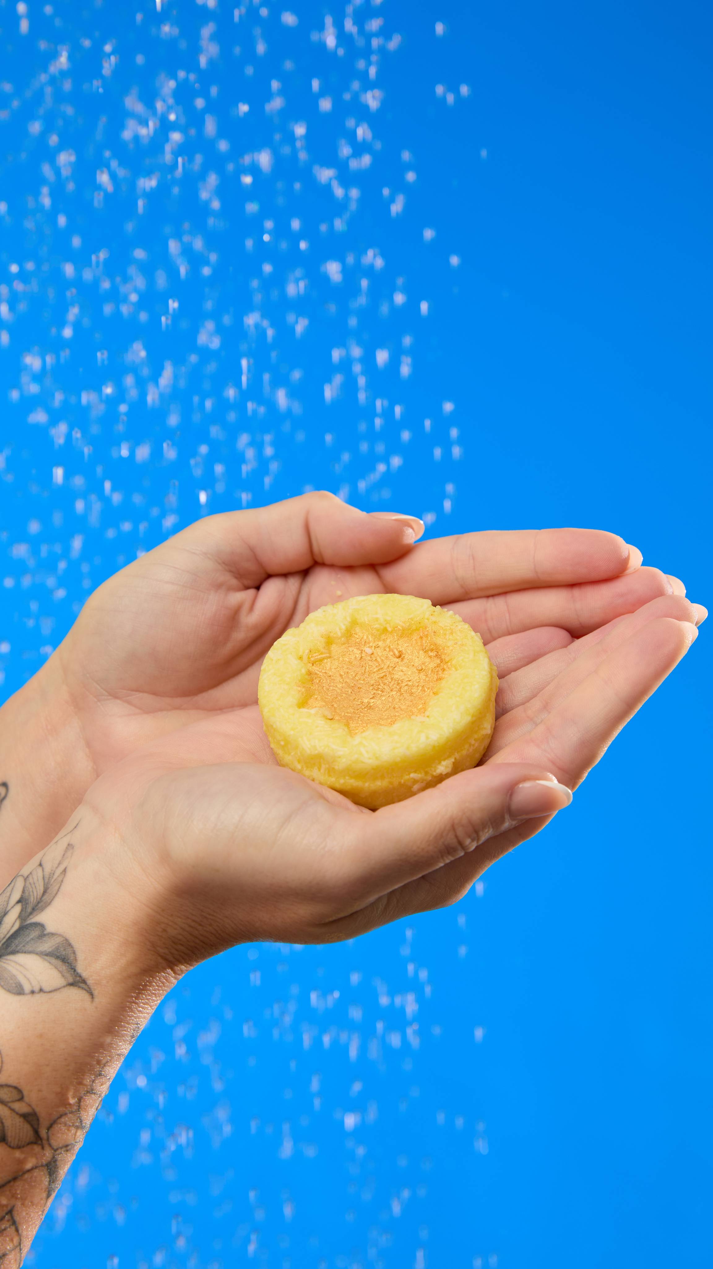 Ray of Hope solid shampoo bar. The image shows hands cupping a pale yellow, round shampoo bar with a central sun-shaped hollow of golden lustre. Water droplets fall on the hands and product against a vibrant blue background.