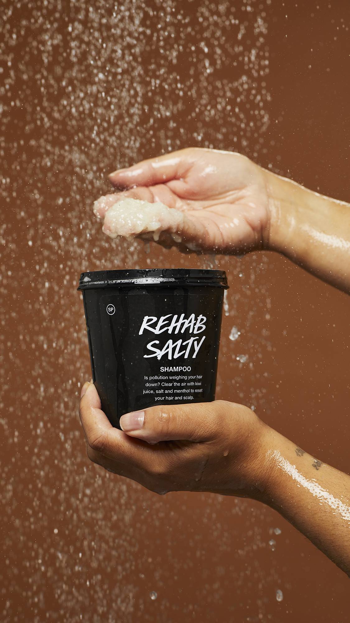 A Close-up image of the Rehab Salty in a LUSH black pot under falling shower water as the model scoops out the product.
