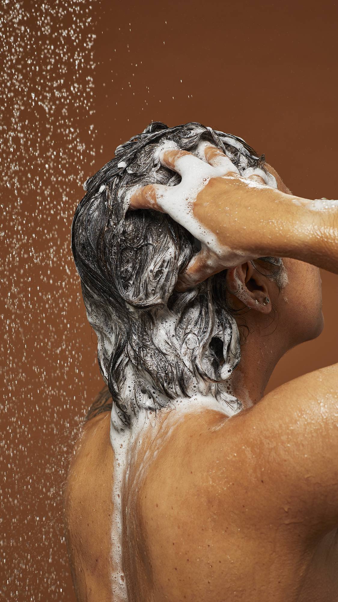 Model is standing under shower water lathering up the shampoo as it falls down their back on a deep earth-tone background.