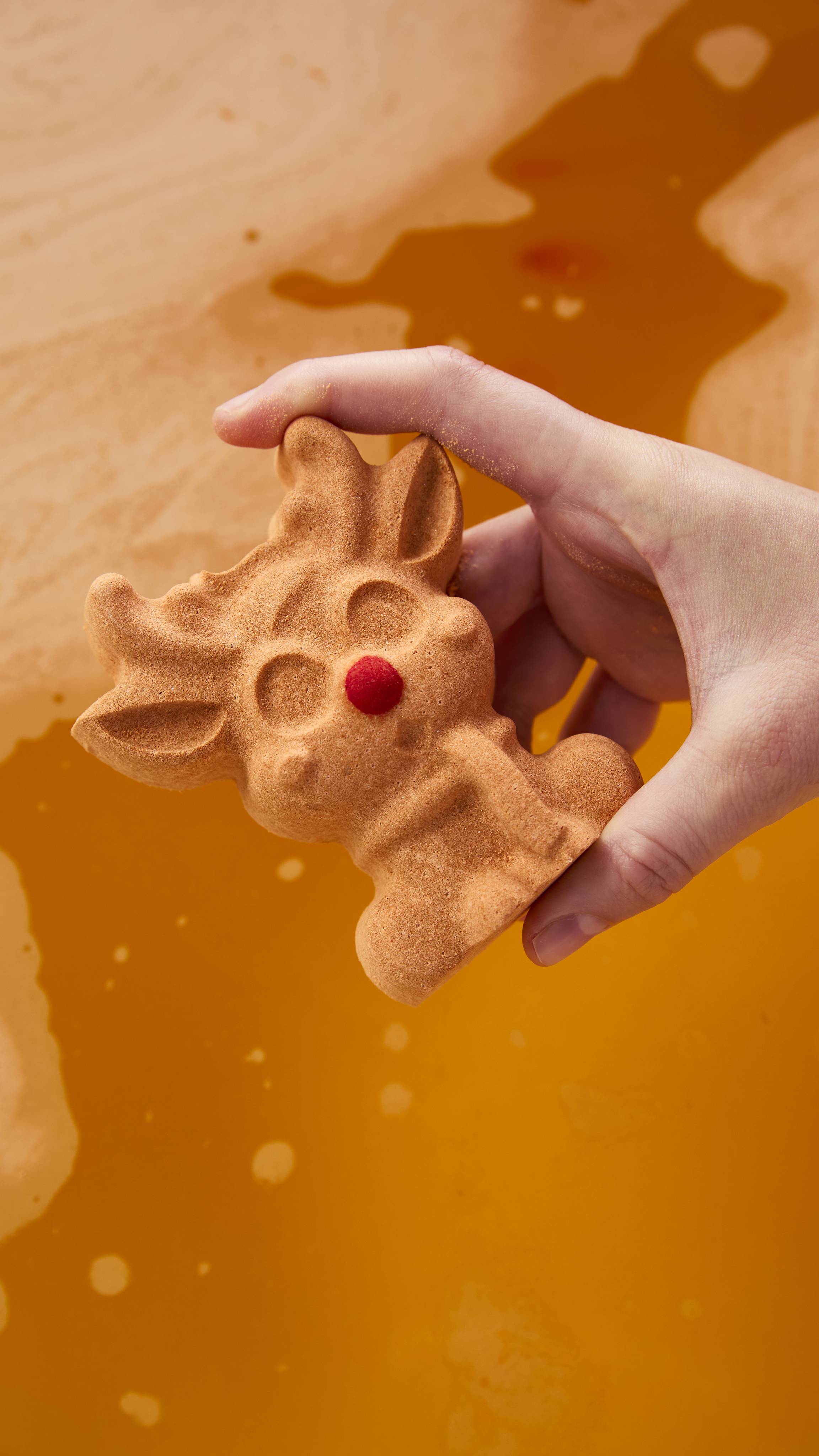 A hand holds a light brown reindeer-shaped bath bomb with a bright red nose over swirling, warm orange bathwater.
