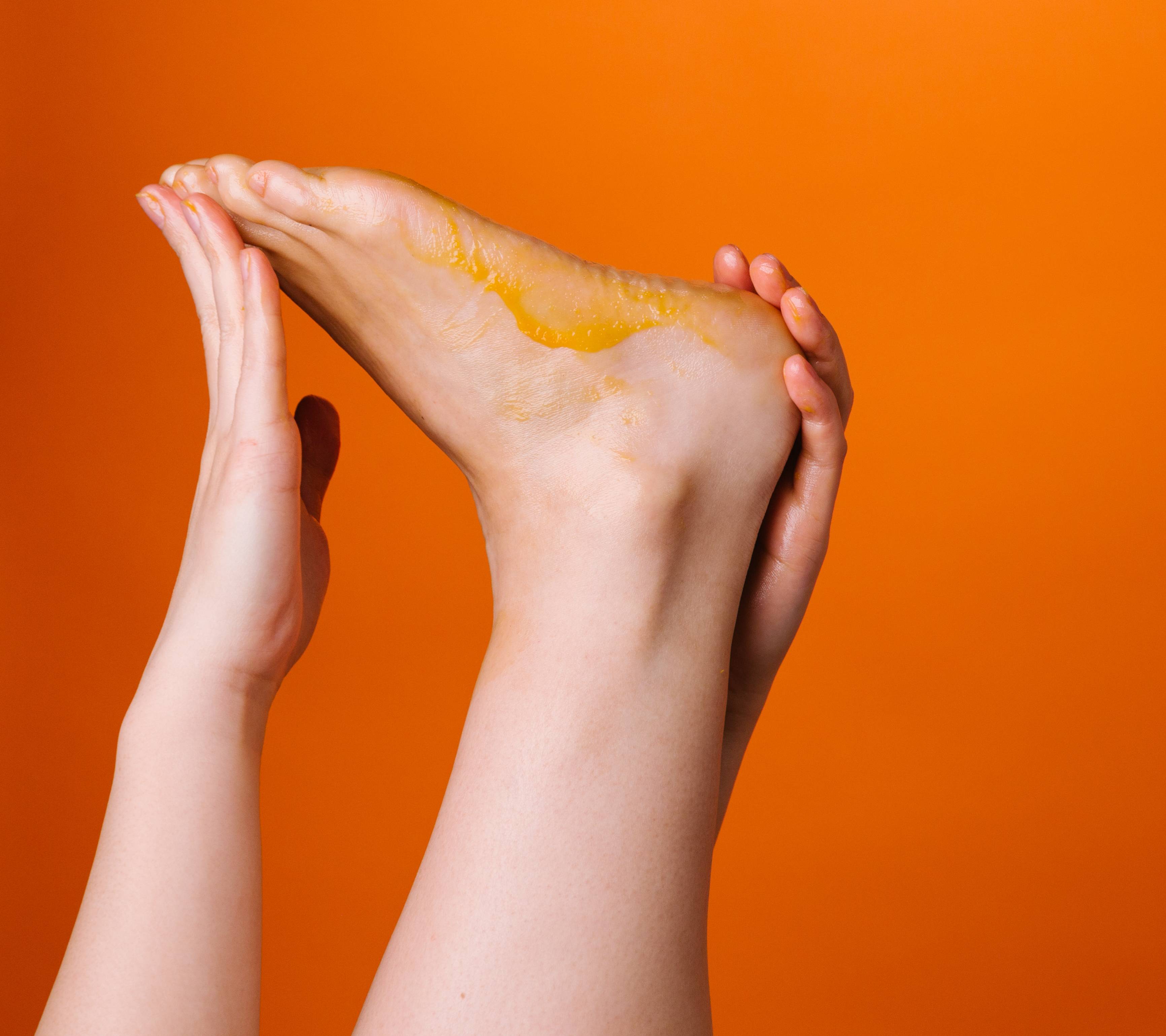 In front of an orange backdrop, a foot is being stretched by two hands, with its sole facing upwards, coated in an orange balm.