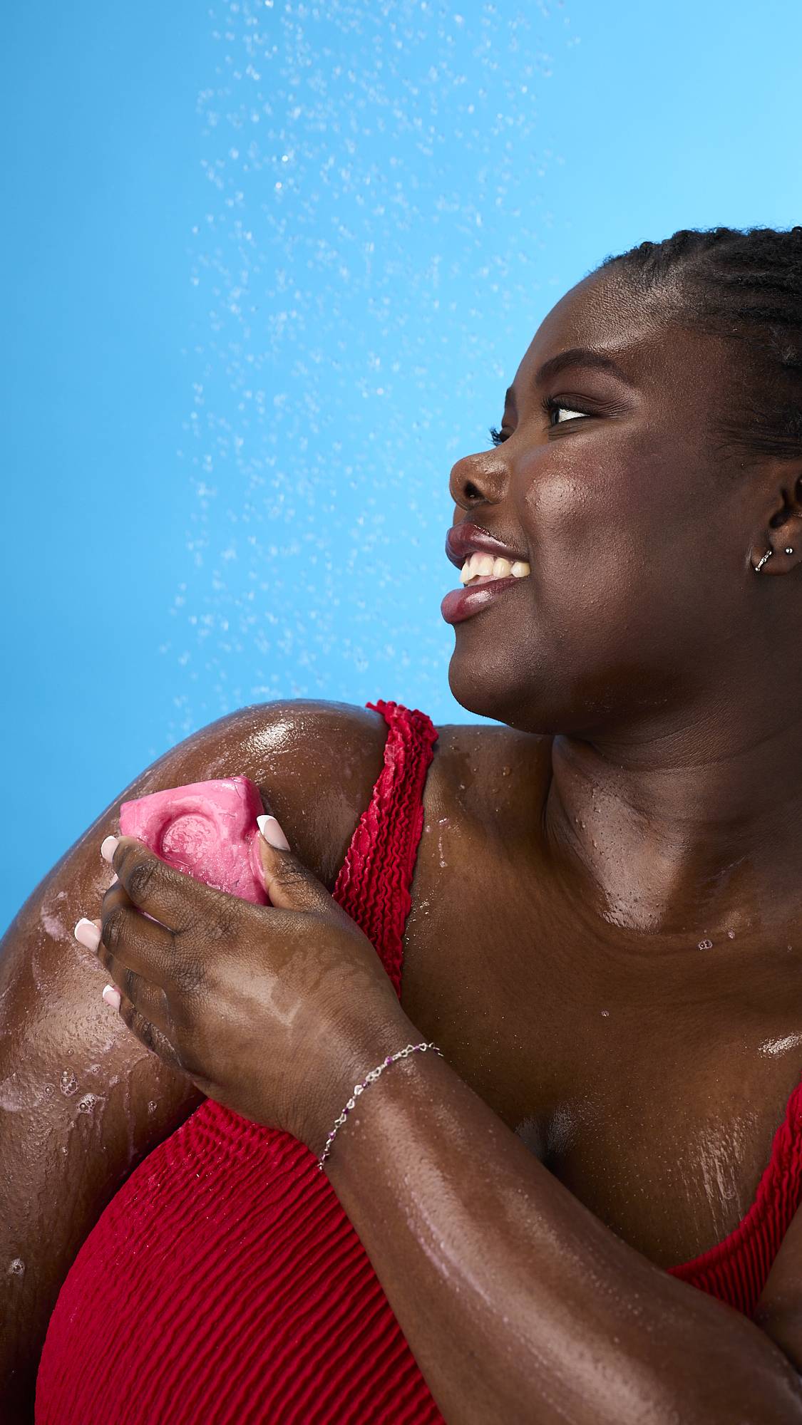 A woman smiles in a shower while using a pink, life-vest-shaped body wash bar on her shoulder. She wears a red swimsuit against a bright blue background, with water droplets and a light lather visible on her skin.