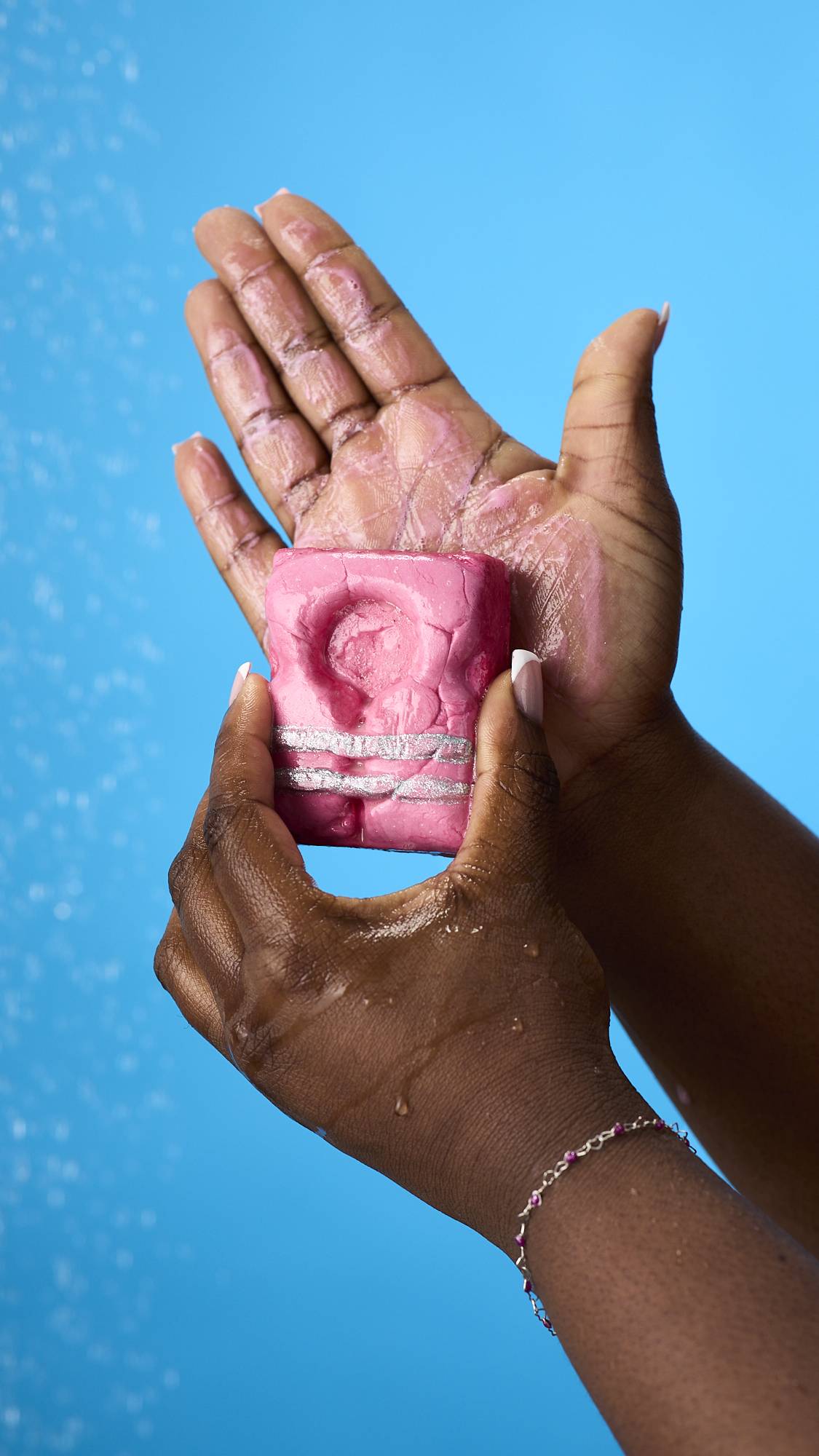 A woman in a red swimsuit smiles in a shower while using a pink, life-vest-shaped body wash bar on her shoulder. Her skin is wet with a light lather against a bright blue background, under a spray of falling water.