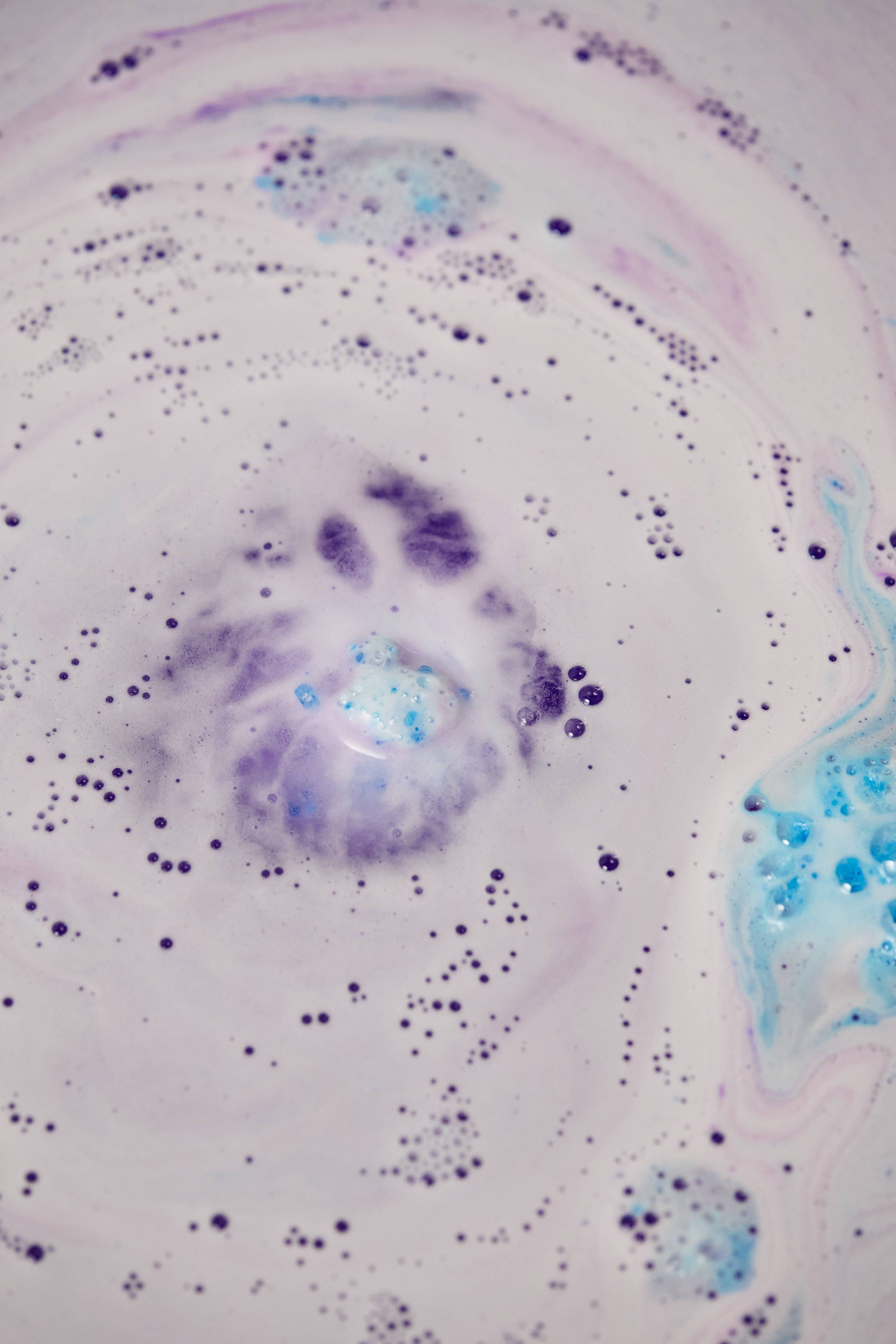 An overhead shot of a Lush "Resilient" bath bomb dissolving into creamy, pale purple water. Concentric swirls of tiny violet bubbles expand outwards from a central purple cloud, while patches of bright turquoise foam float nearby.