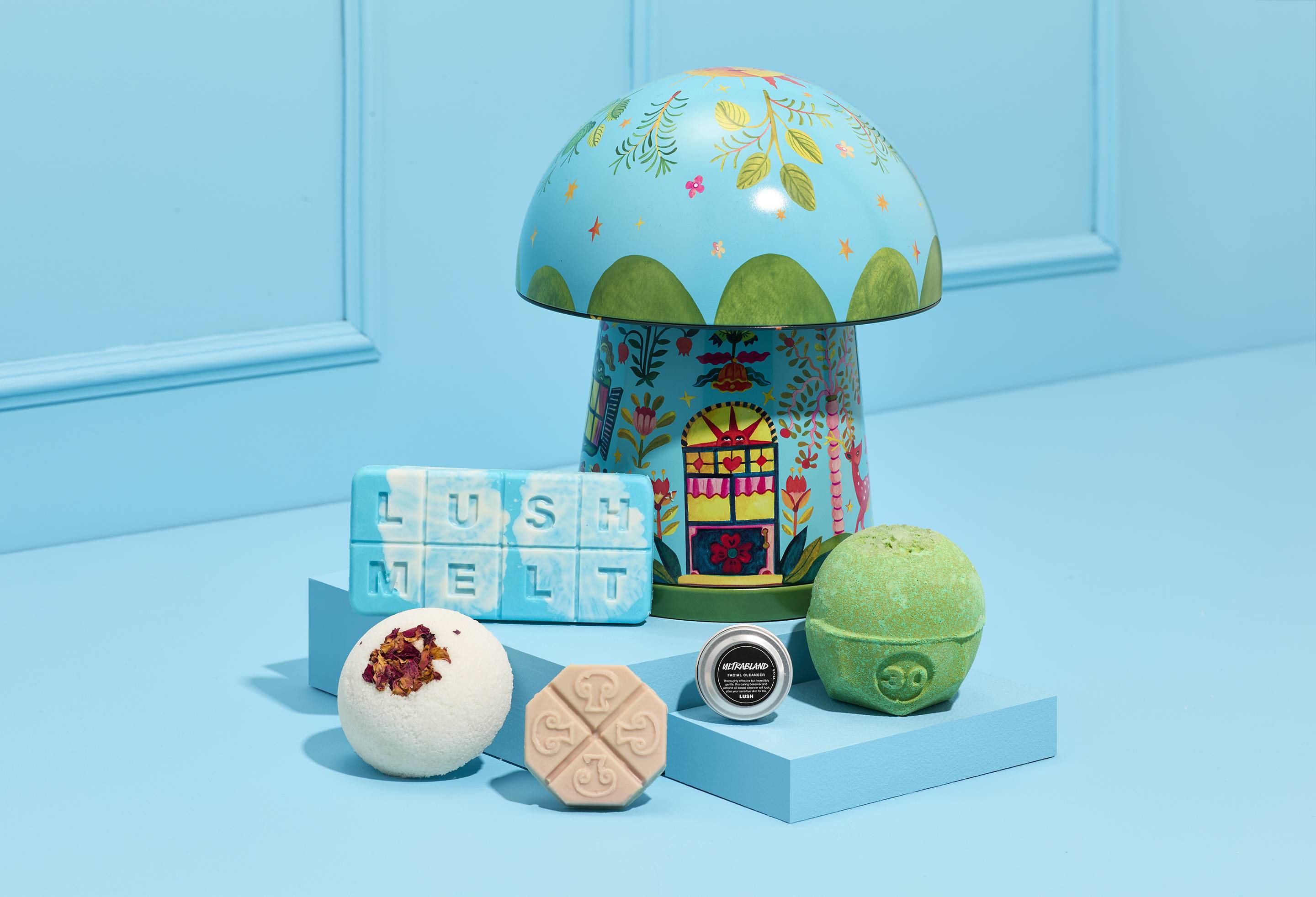The image shows the mushroom-shaped "Rewild" gift tin with a blue, folk-art-pattern, sitting on a blue tiered platform. Arranged in front is a Lush Melt bar, a bath bomb, a solid facial serum bar, a tin of facial cleanser and a solid body scrub.