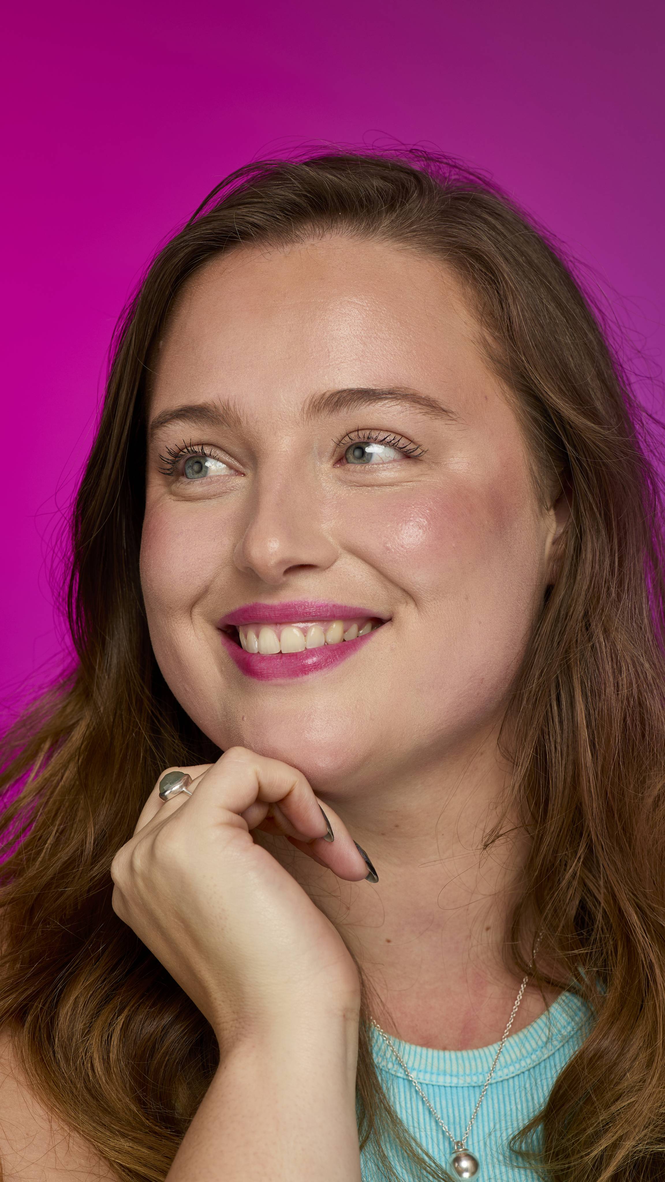 A person with long brown hair, wearing a ribbed blue top, smiles with a vibrant magenta Rhubarby~ Lip Stain against a bright pink background. A silver ring and a pearl necklace are visible.