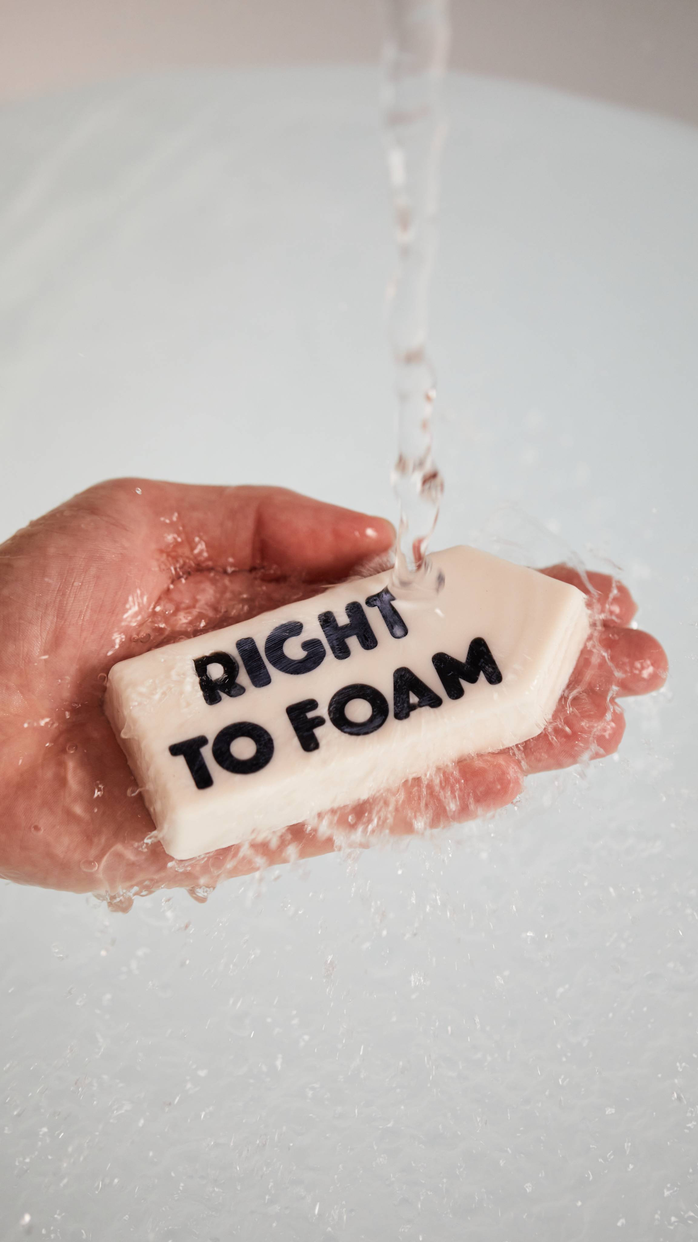 A close-up of the model's hand as they hold the white, sign-shaped, Right to Foam soap under running water.