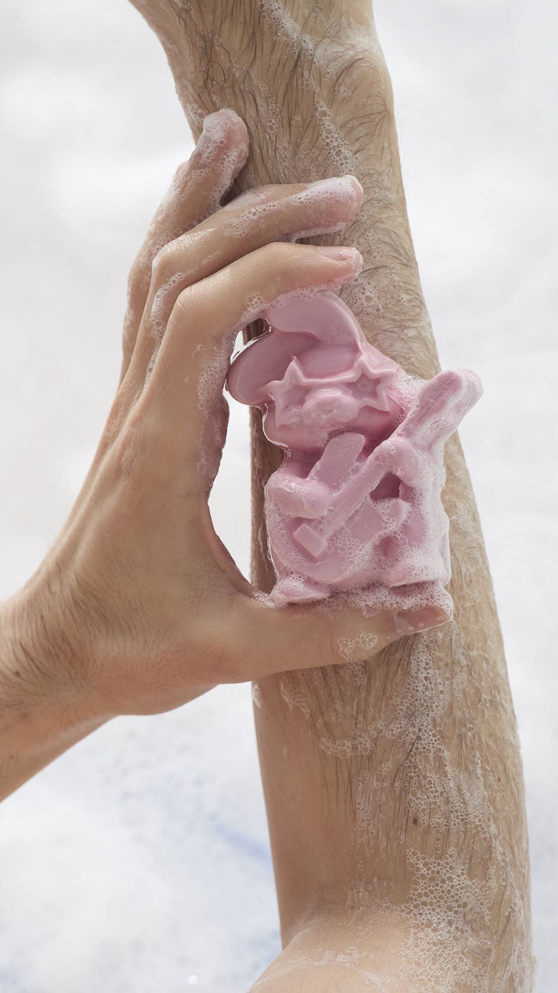 A close-up of a pink rabbit-shaped soap being used to lather a person's arm. The bunny, wearing star sunglasses and holding a guitar, is covered in creamy white suds against the skin, with a bubbly bath in the background.