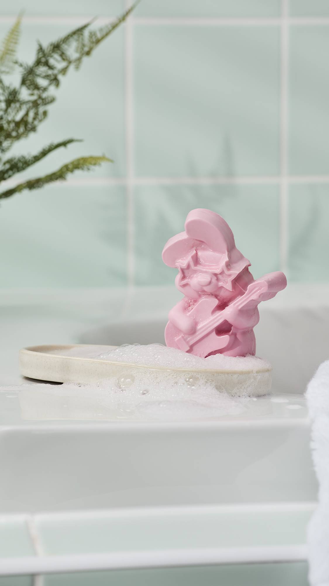 A pink rabbit-shaped soap wearing star-shaped sunglasses and playing a guitar. It sits on a white soap dish with fluffy white bubbles, set against a background of light mint green tiles and a small green plant.