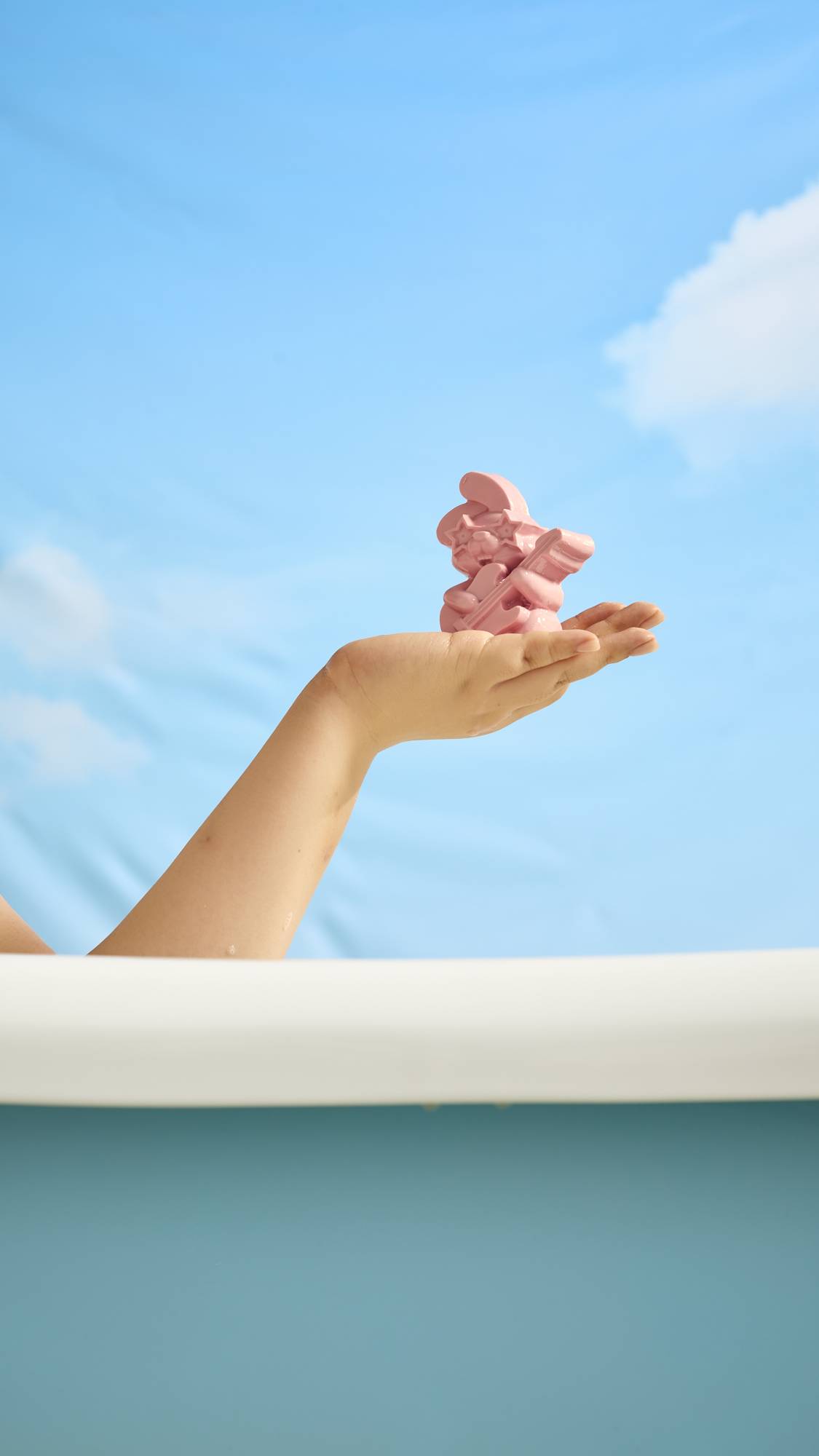 A close-up image of the model's arm over the side of an outdoor bath under blue skies as they gently hold the Rockstar Rabbit soap in their palm. 