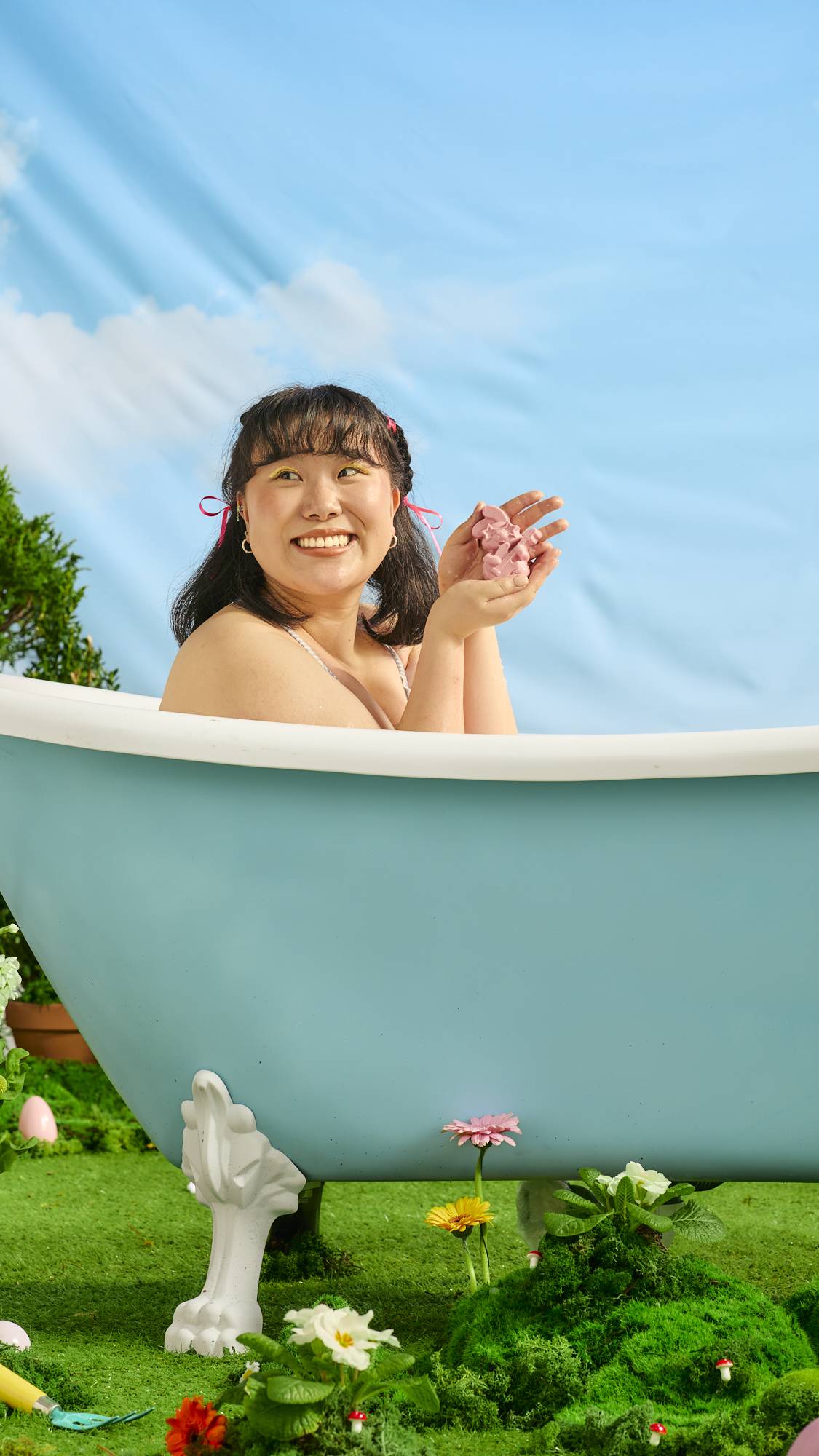 The model is in an outdoor bath under blue skies, surrounded by glass and spring flowers as they smile and hold the Rockstar Rabbit soap with both hands. 