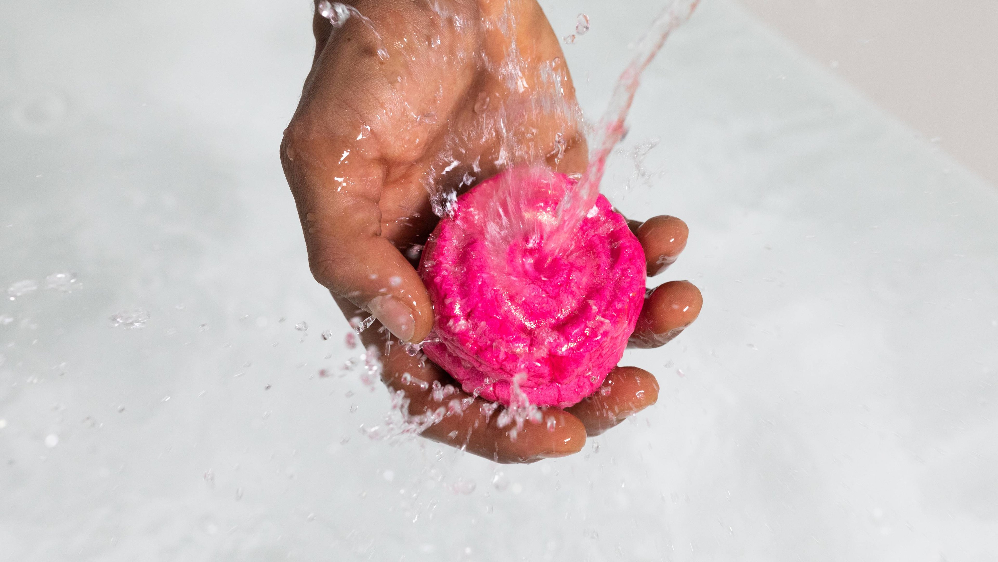The model holds the sparkly, pink Rose Jam Bubbleroon bubble bar under running water as it flows to create bubbles below.