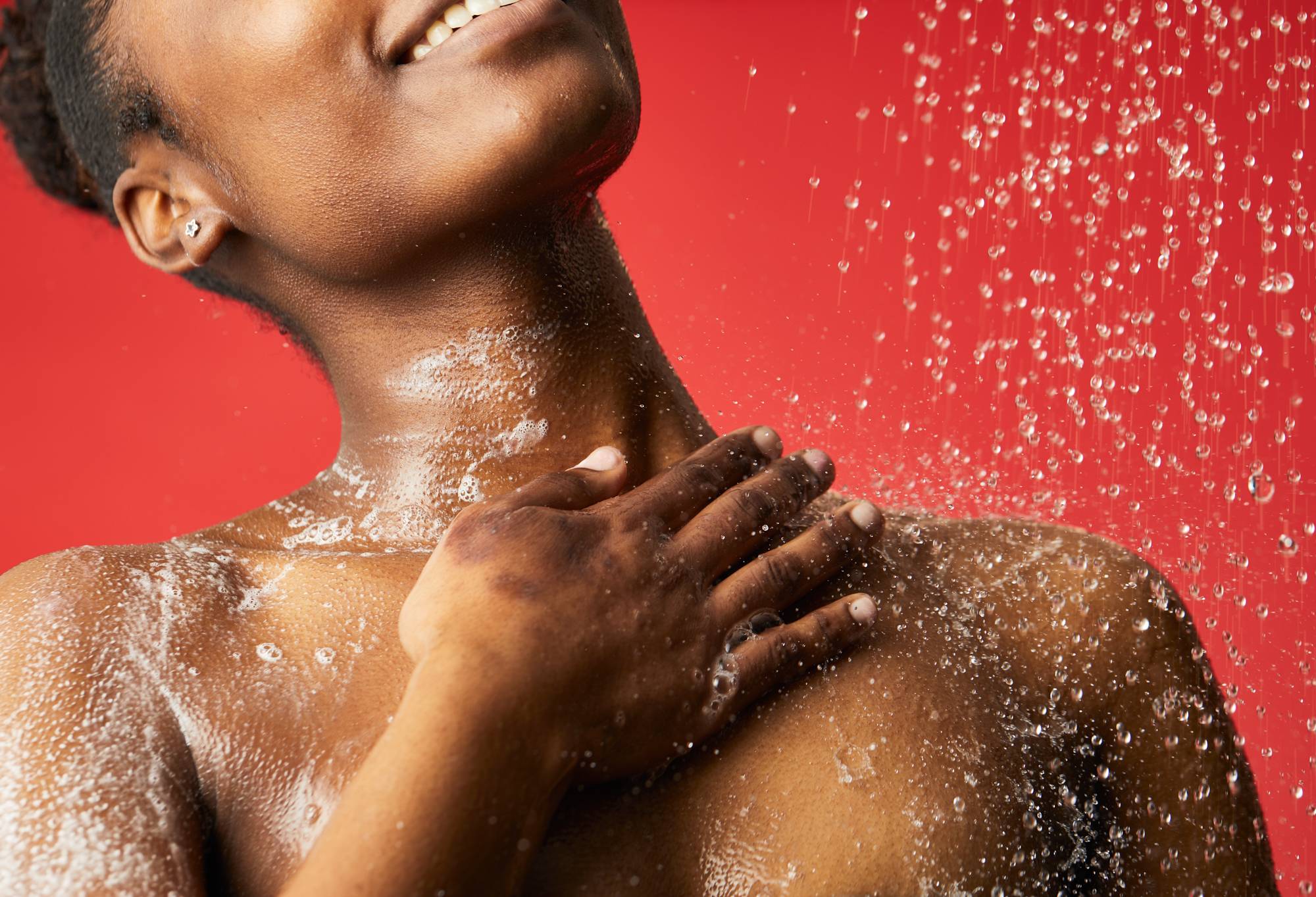 The shower gel is lathered up over a model's right shoulder with their hand, in front of a light red background.
