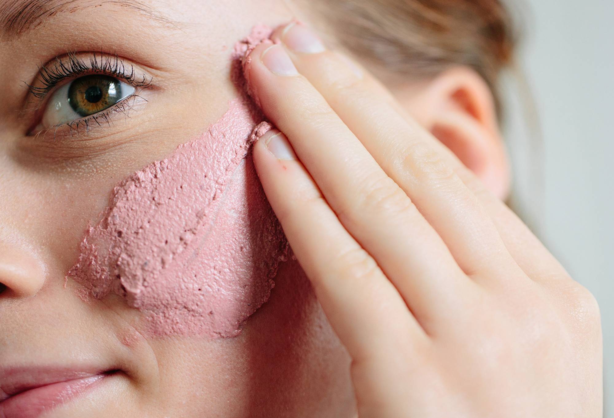 Thick, rosy pink Rosy Cheeks face mask is applied upwards to the cheeks of a softly smiling face.