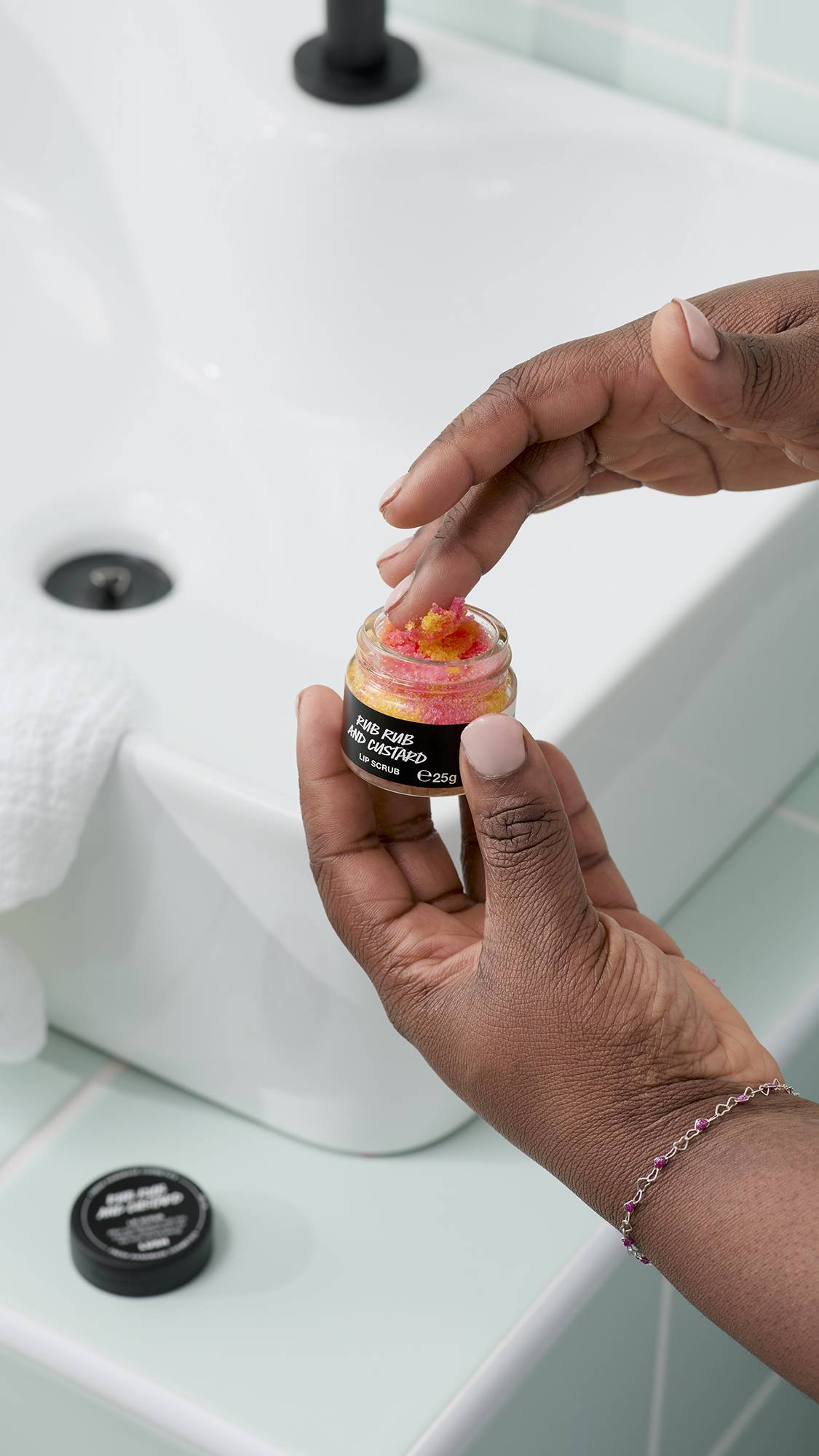 The image shows a model's hands holding an open small glass jar of Lush 'Rhubarb and Custard' lip scrub, which contains bright pink and yellow sugar crystals. The background shows a white bathroom sink and light blue tiles.