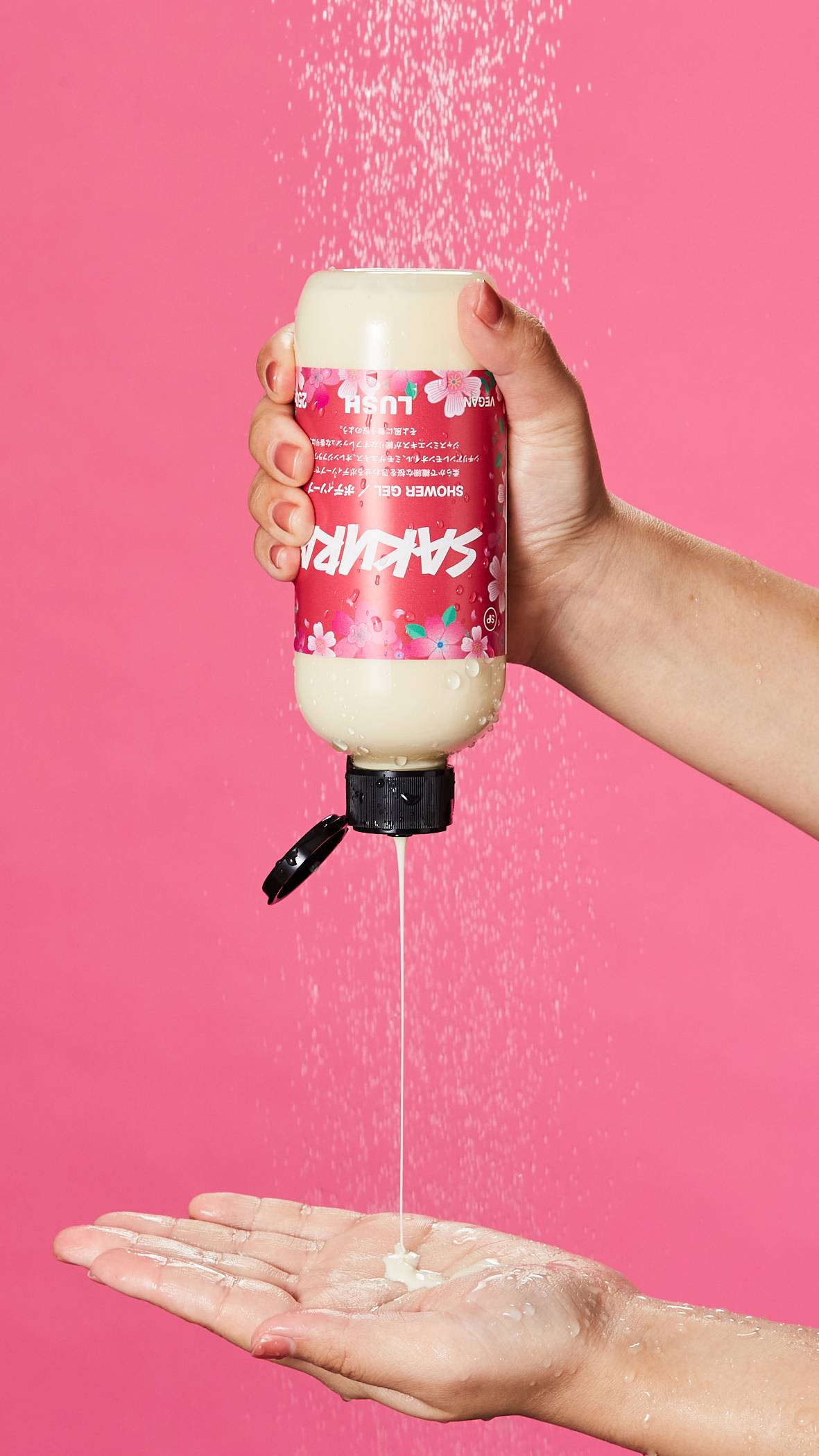 A close-up of the model squeezing the Sakura shower gel from the bottle into their palm under running shower water with a cherry blossom-pink background. 