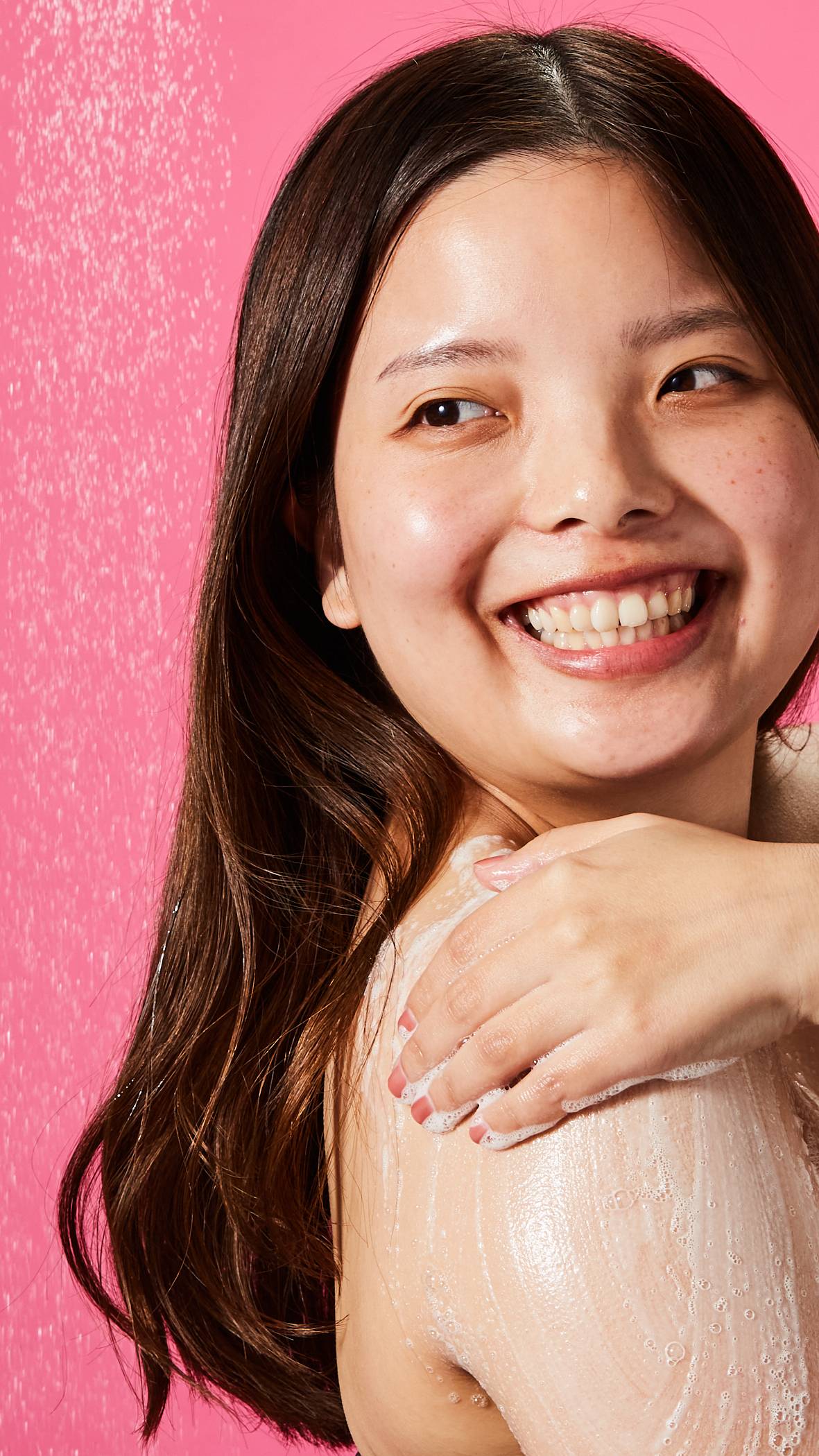 The model is smiling and looking over their shoulder as they stand under the shower and lather up their upper arm with Sakura shower gel on a pink background. 