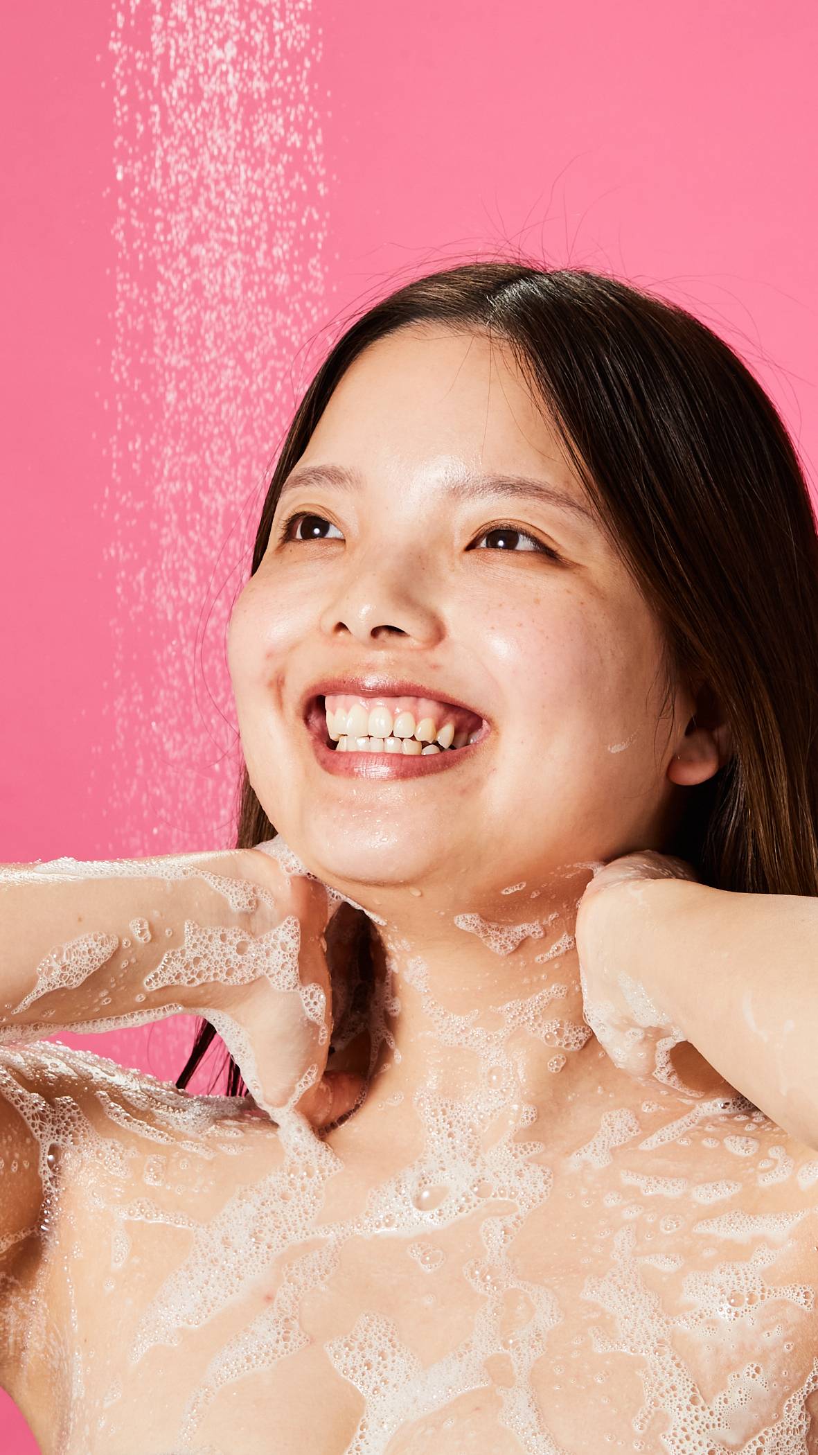 The model is standing under running shower water on a vibrant, pink background as they lather up their neck and chest with the Sakura shower gel. 